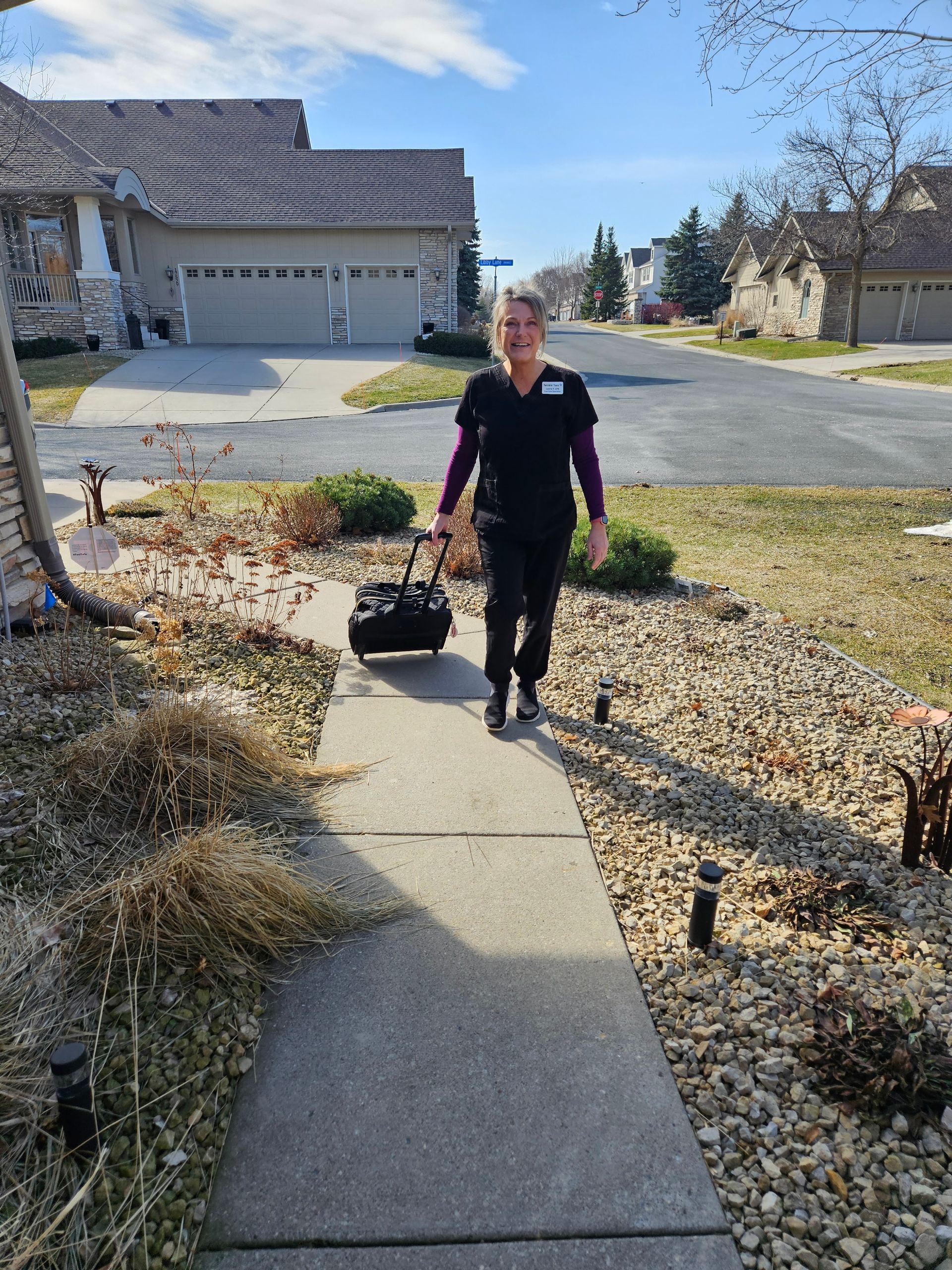 Laurie, Twinkle Toes, in black medical scrubs, walking up a residential sidewalk, pulling a small black bag.