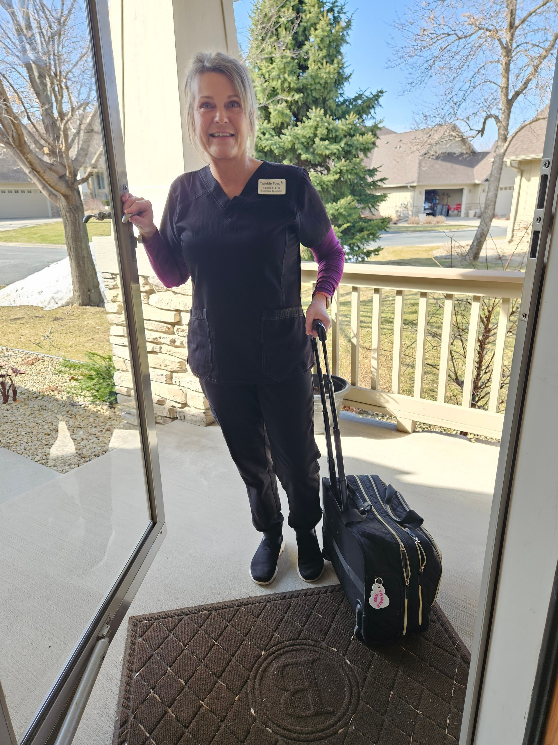 Laurie, Twinkle Toes, in black scrubs, stands in a doorway, as seen from inside a house on a sunny day.