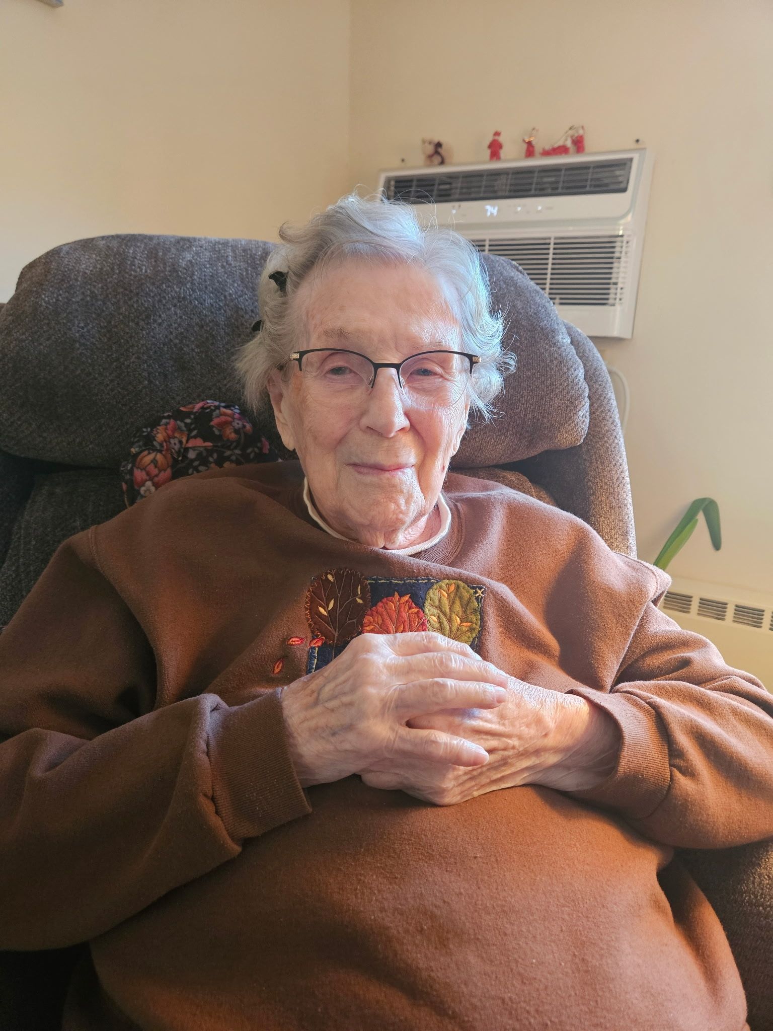 Elderly woman wearing glasses, sits in a recliner, hands clasped. Brown sweater, indoor setting.