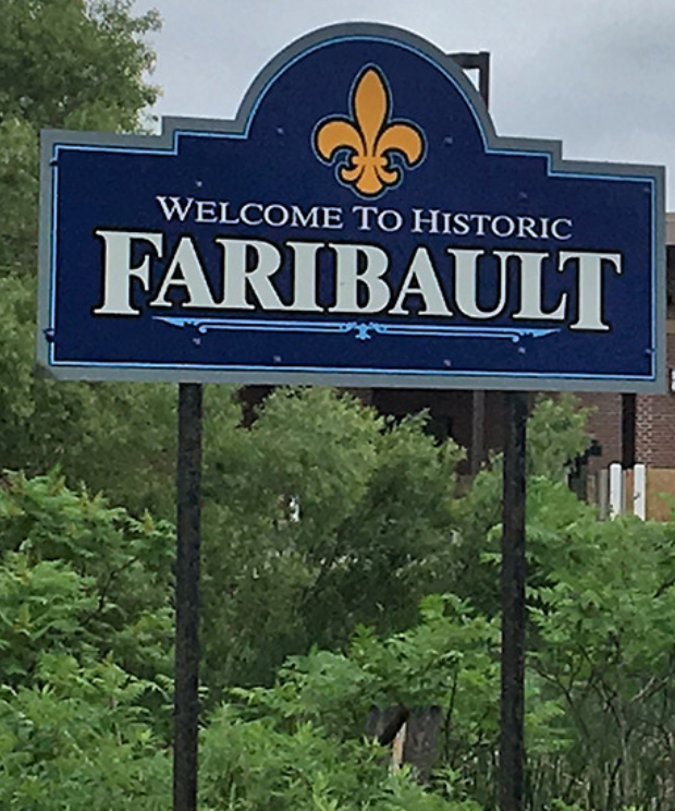 Blue “Welcome to Historic Faribault” sign with gold fleur-de-lis above green trees