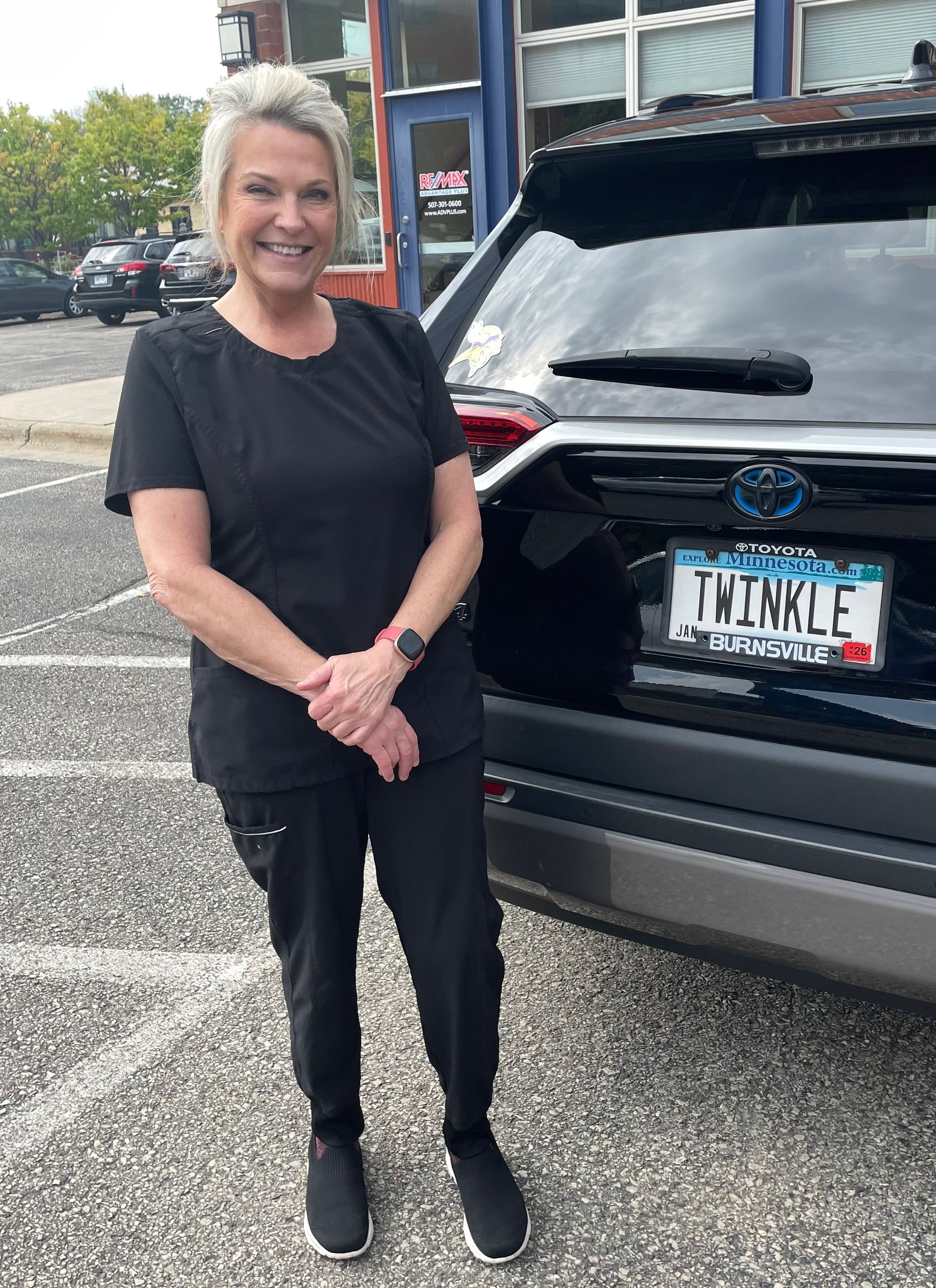 Nurse Laurie from Twinkle Toes, in black scrubs, smiles and stands beside a black SUV with the license plate 