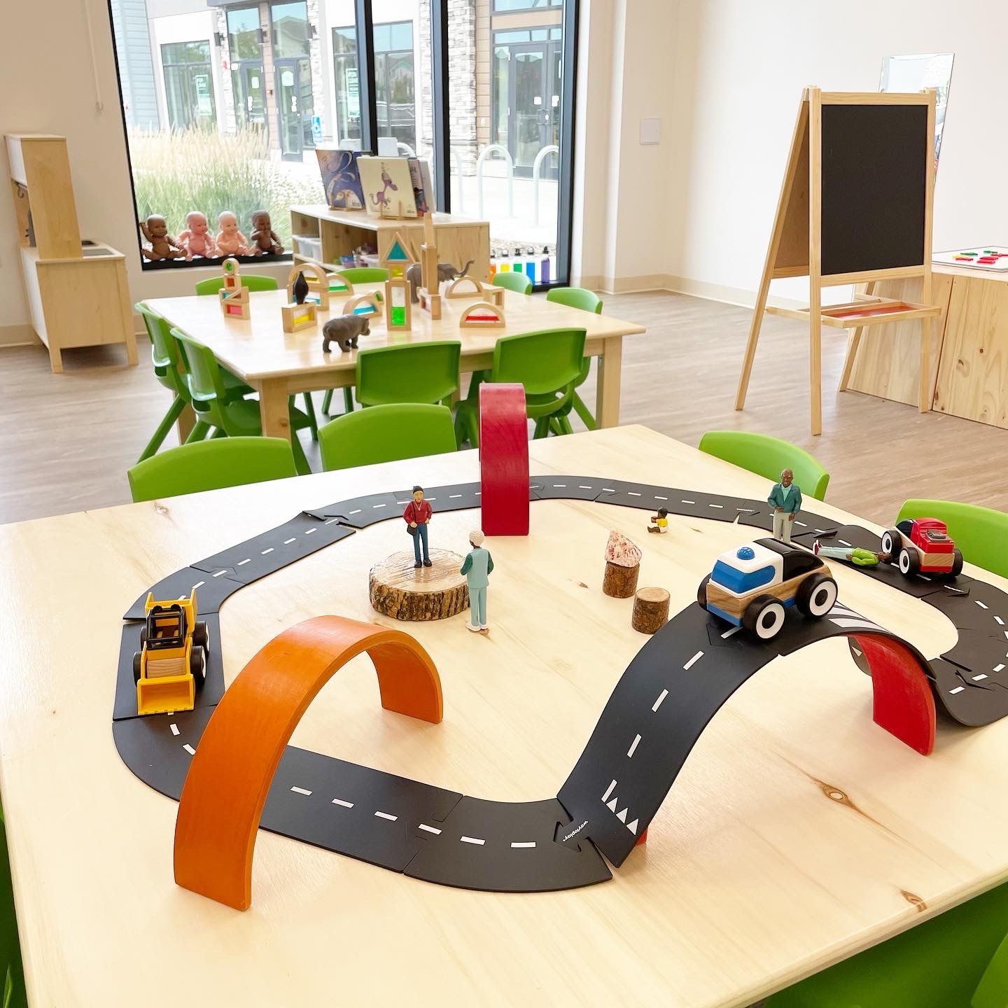 Toy cars on a curved road track in a bright classroom with green chairs and a small chalkboard.