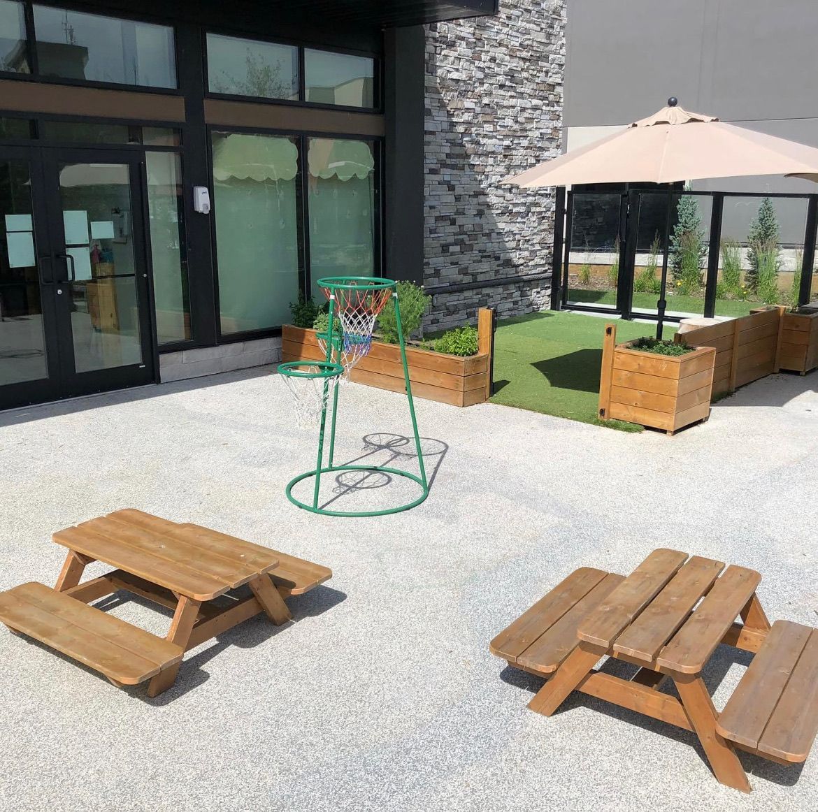 Outdoor patio with picnic tables, planter boxes, a green fire pit, and a beige umbrella beside a building.