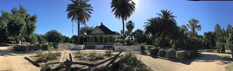 Tree Services West Covina -Panoramic view of a lush garden with a quaint house surrounded by tall palm trees and a white picket fence under a clear blue sky.