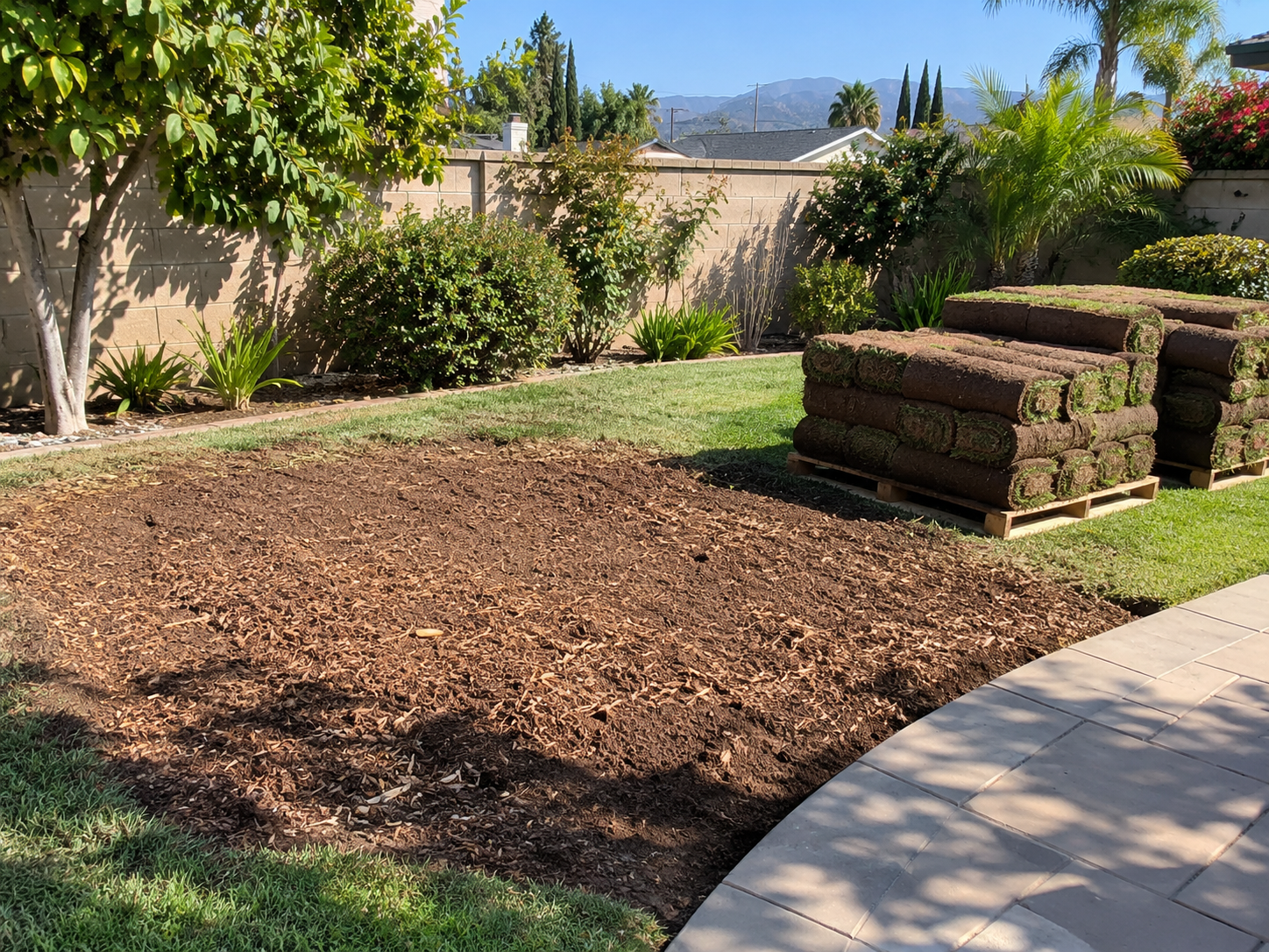 West Covina backyard with freshly ground stump area raked level beside stacked sod rolls and a paved patio edge