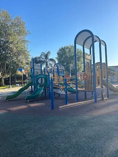 Tree Services West Covina-A sunny playground with multiple blue and yellow slides, climbing structures, and ladders on a rubber floor, surrounded by trees and clear skies.