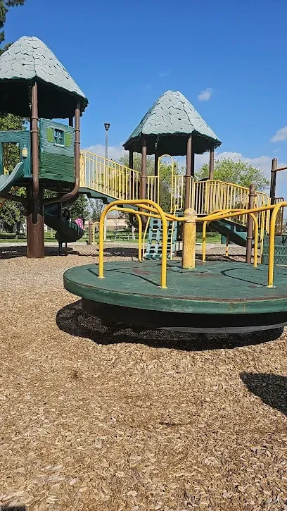 Tree Services West Covina-Children's playground with a wooden structure featuring green roofs, slides, and a bridge. In front, a green merry-go-round with yellow bars. Bright, sunny day.