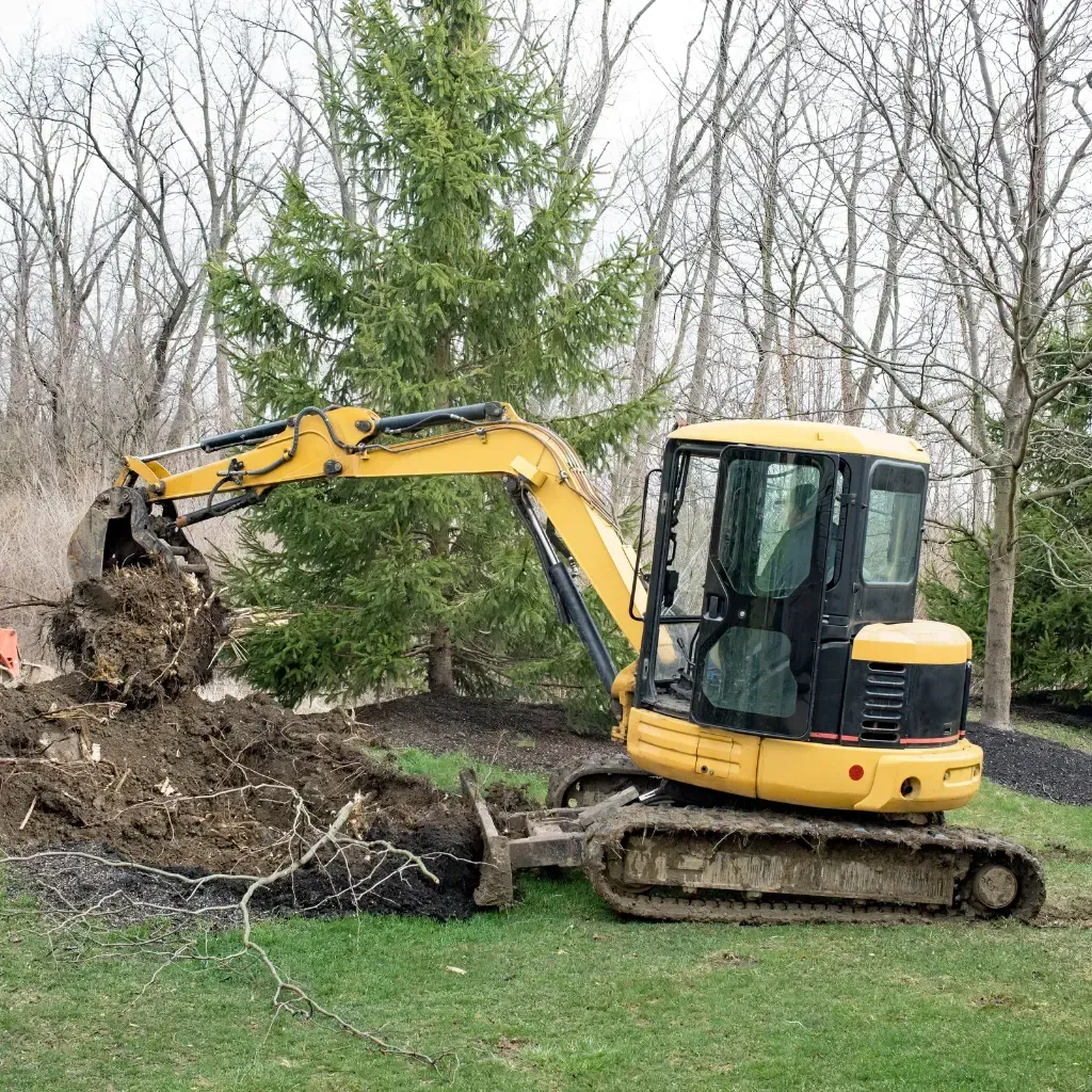 Tree Removal Service West Covina -Yellow excavator digging in a grassy area with dirt and tree branches.
