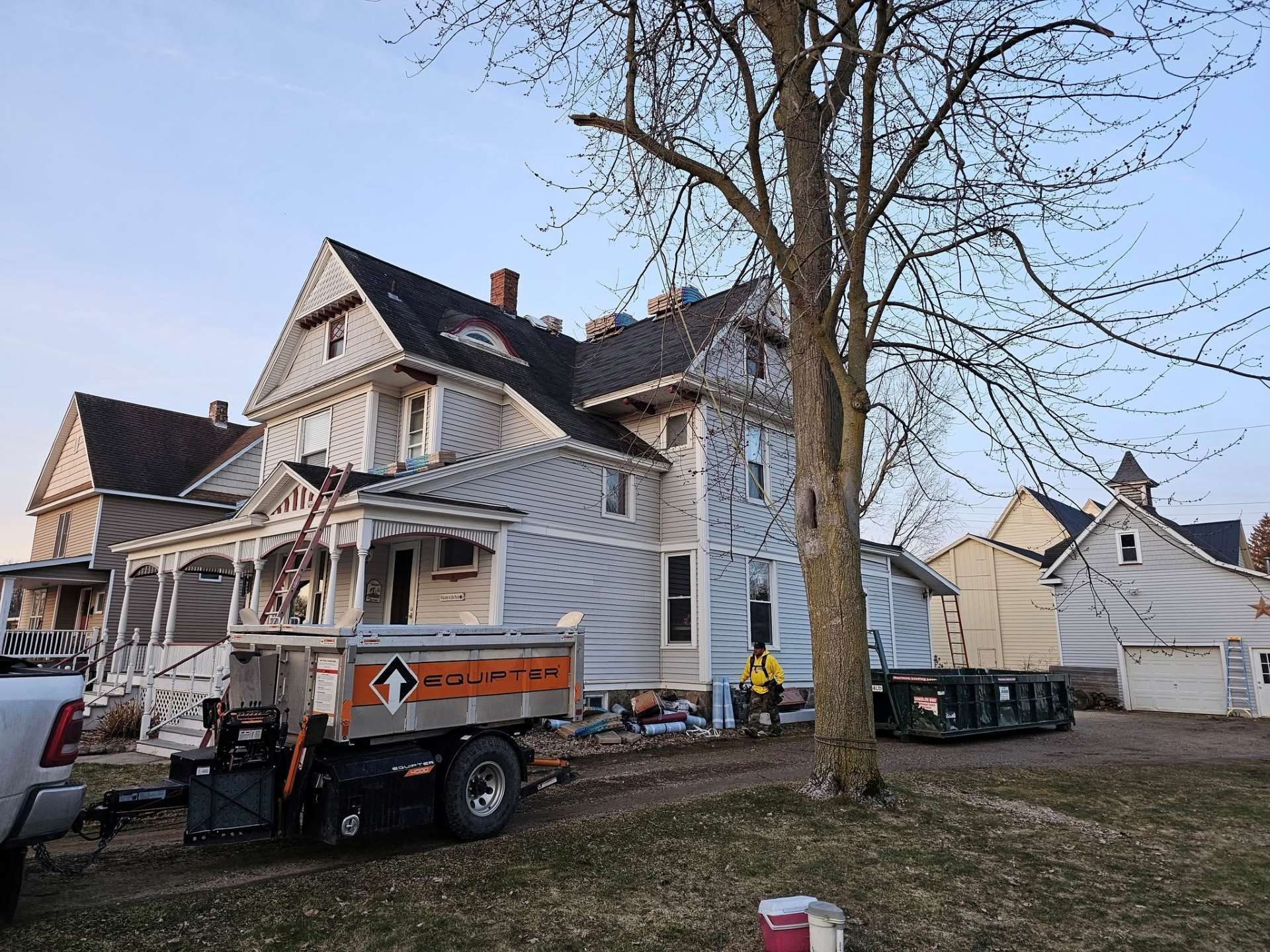 A house under renovation with a dump trailer in the front yard, ladders against the porch, and a worker nearby.