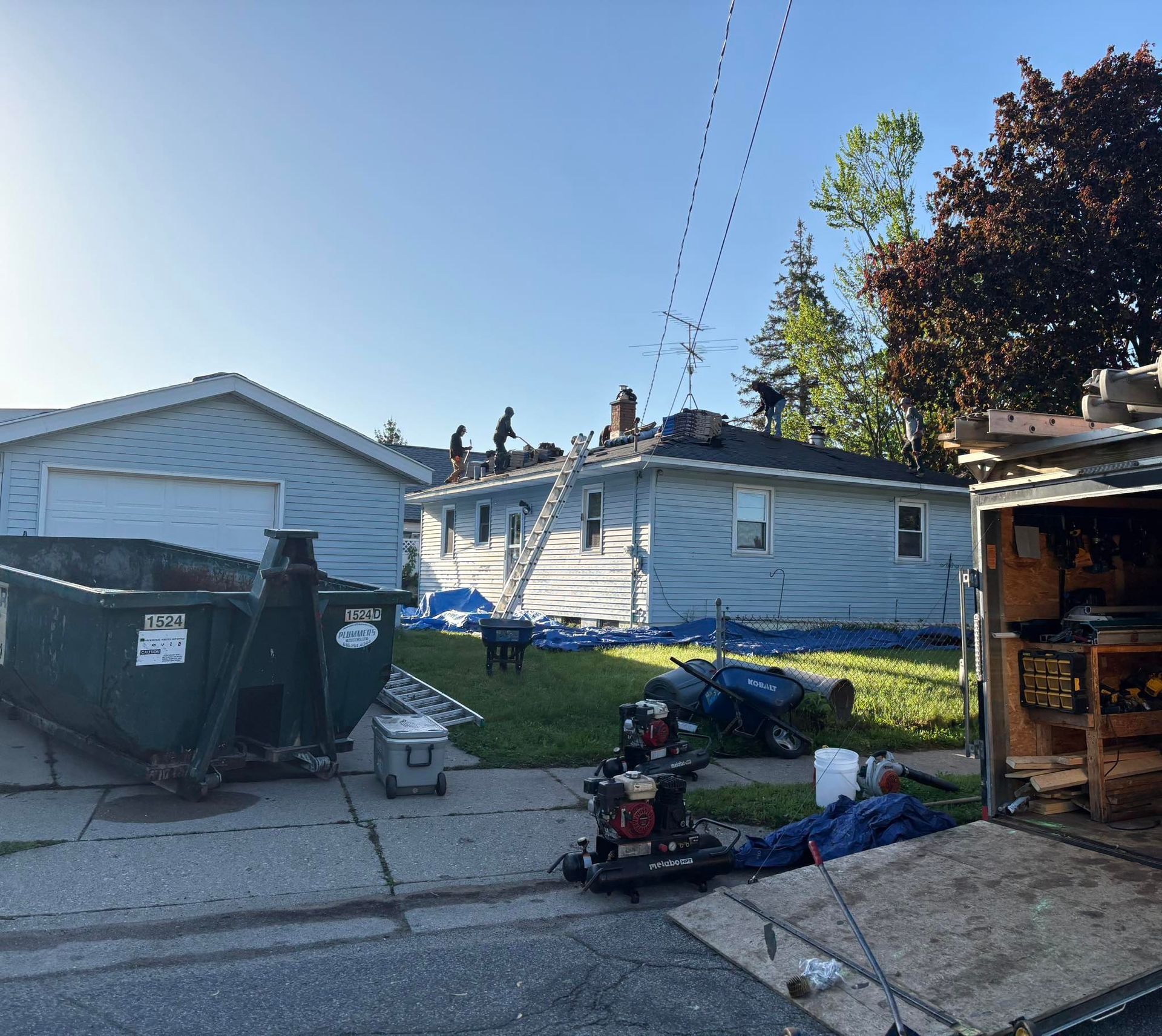 A crew works on replacing the roof of a light blue house, with a large dumpster and work trailer in the driveway.