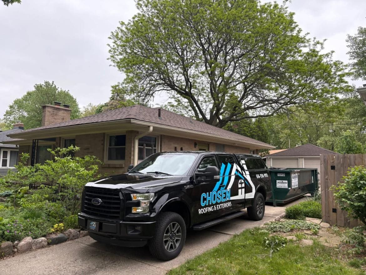 A black Chosen Roofing & Construction pickup truck parked in the driveway of a house with a dumpster nearby.