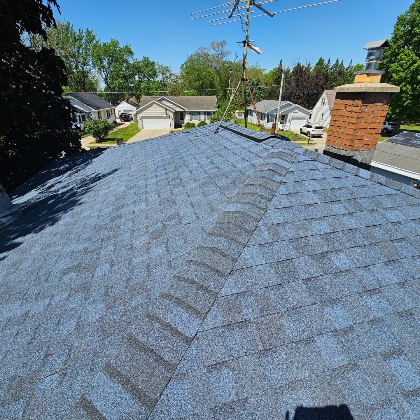 A high-angle view of a residential blue shingle roof with a brick chimney and neighborhood houses in the background.