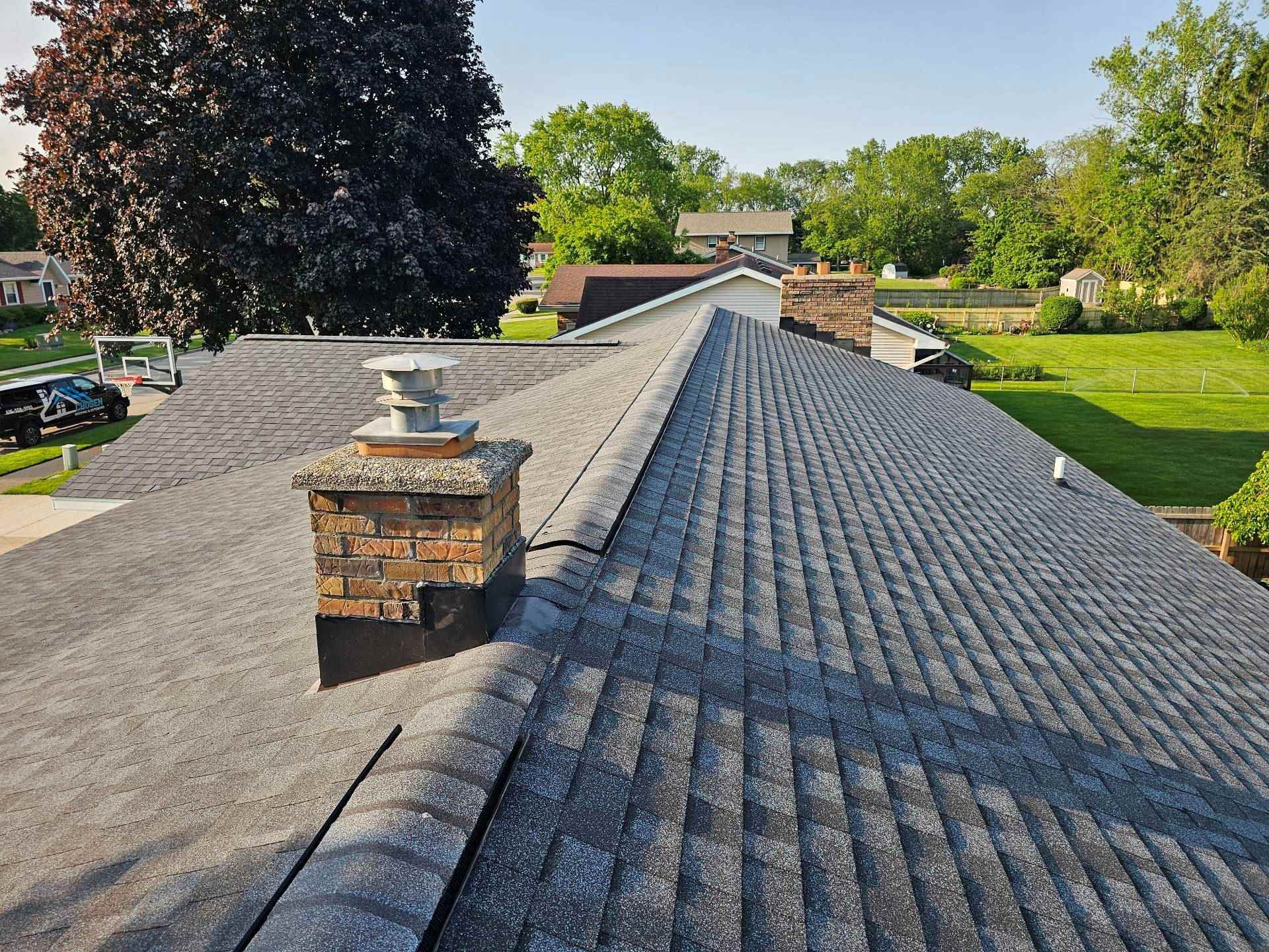 A high-angle view of a gray asphalt shingle roof featuring a brick chimney with a metal vent cap on a sunny day.