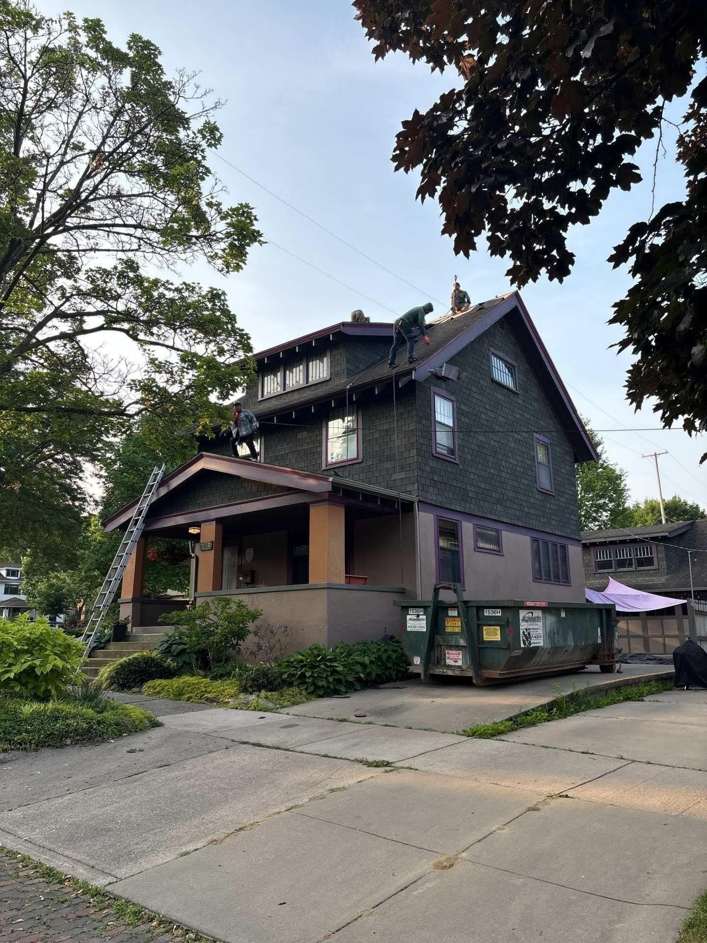Workers are on the roof of a dark-shingled two-story house with a dumpster in the driveway.