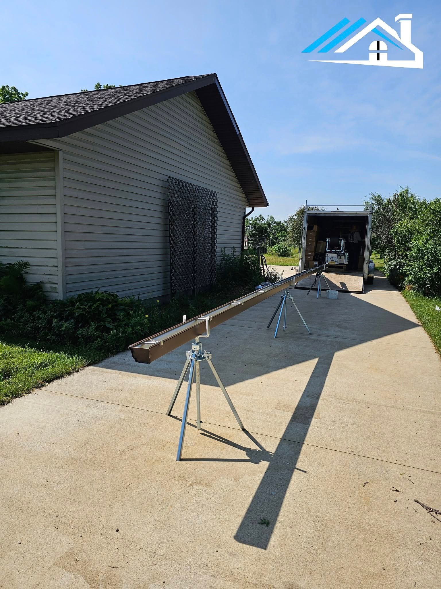 A long brown gutter section rests on two tripod stands on a concrete driveway outside a house.