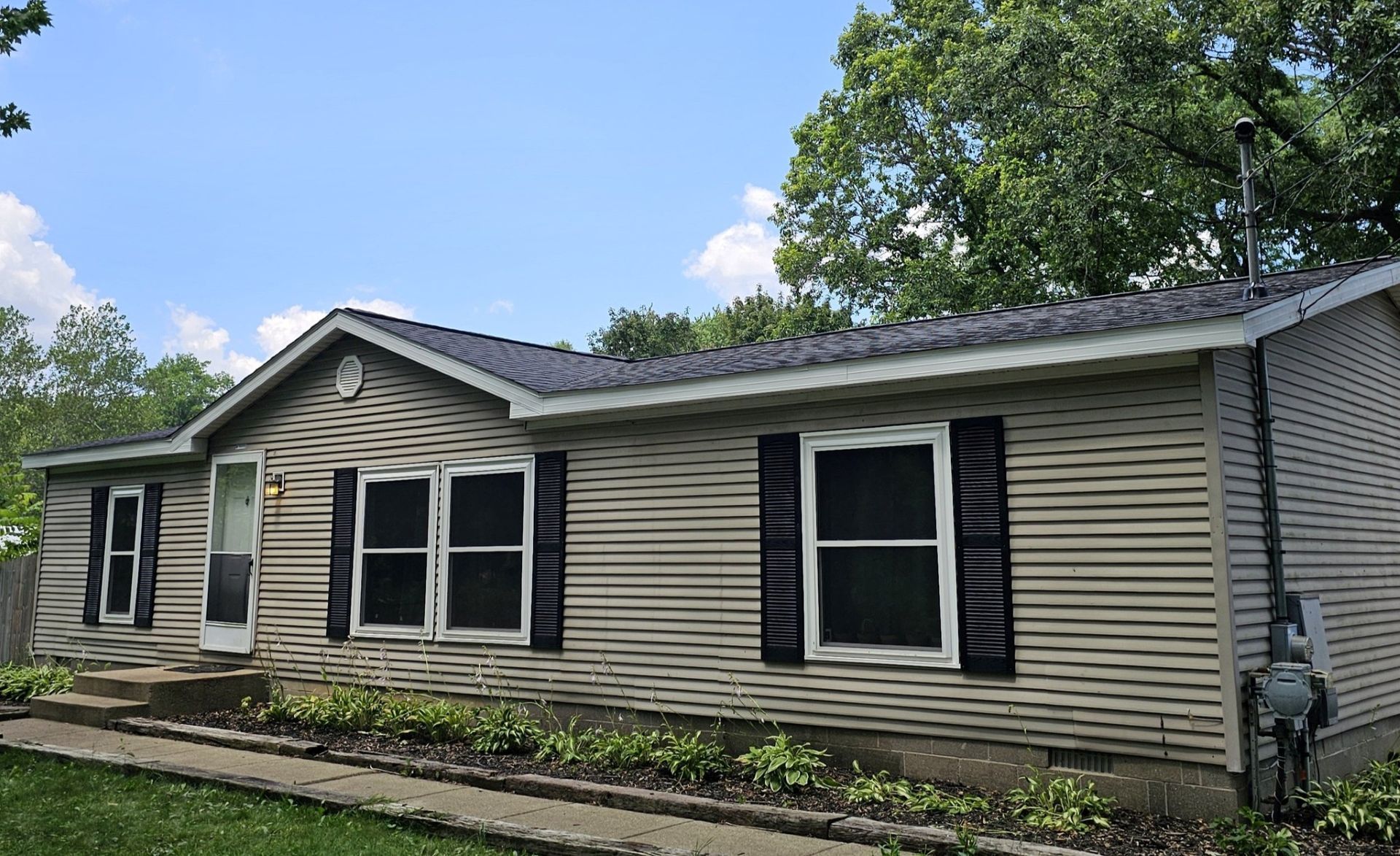 A single-story house with beige horizontal siding, black shutters, and a dark shingled roof under a bright blue sky.