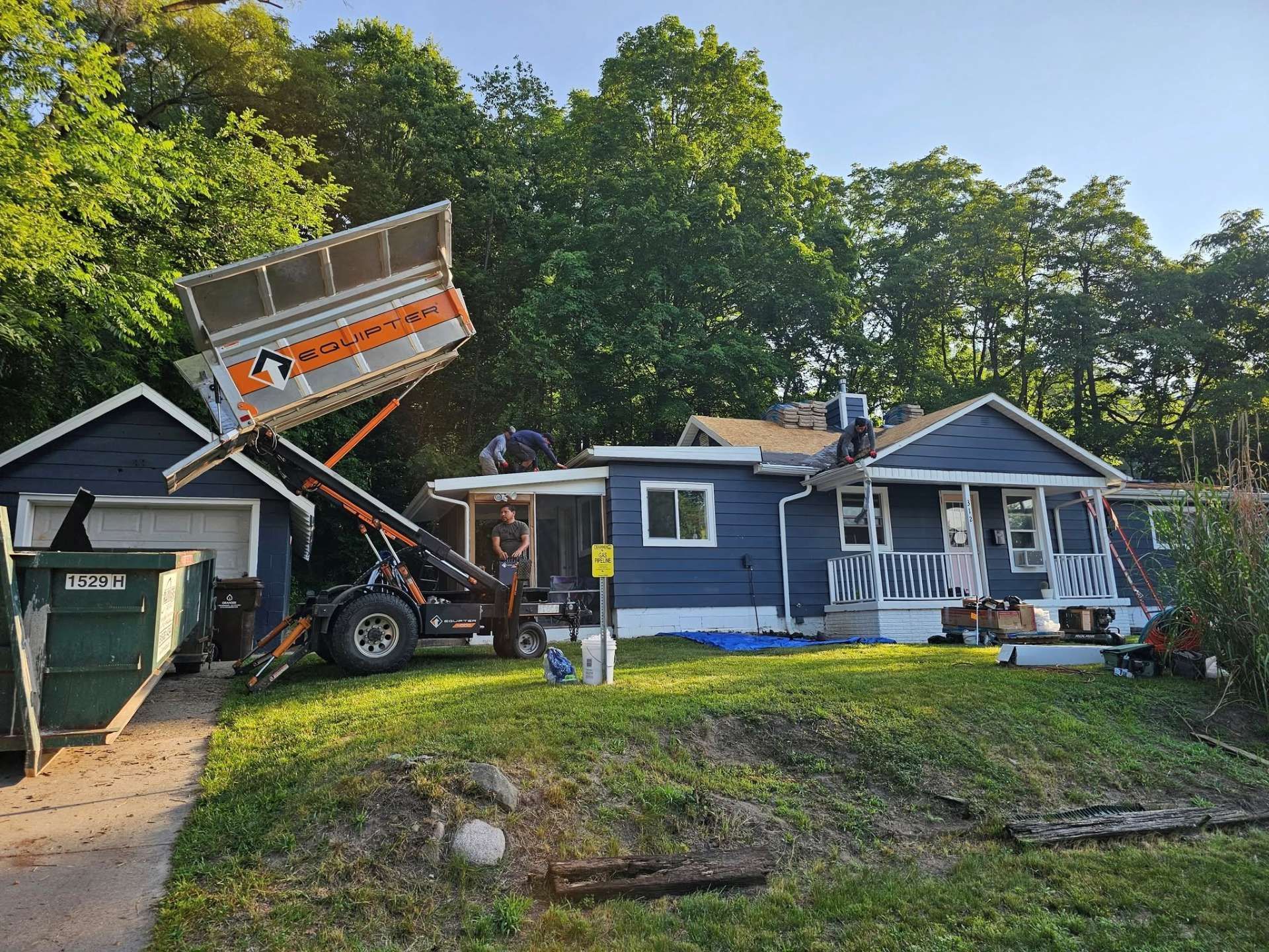 A blue house undergoing a roof replacement with a dumpster trailer lifted high by a piece of construction equipment.