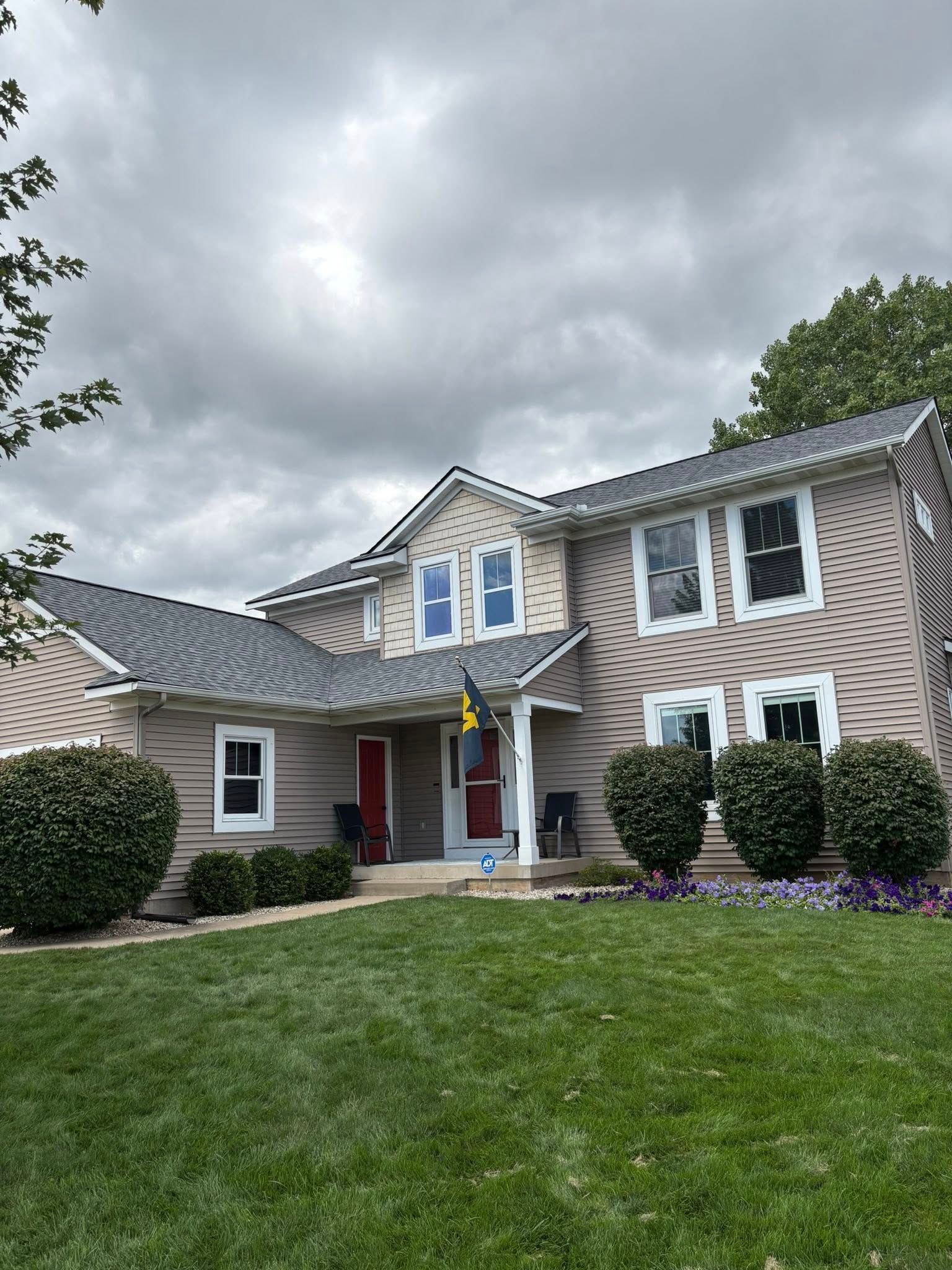 A two-story tan suburban house with a gray roof and dark trim, surrounded by a green lawn and neatly trimmed bushes.
