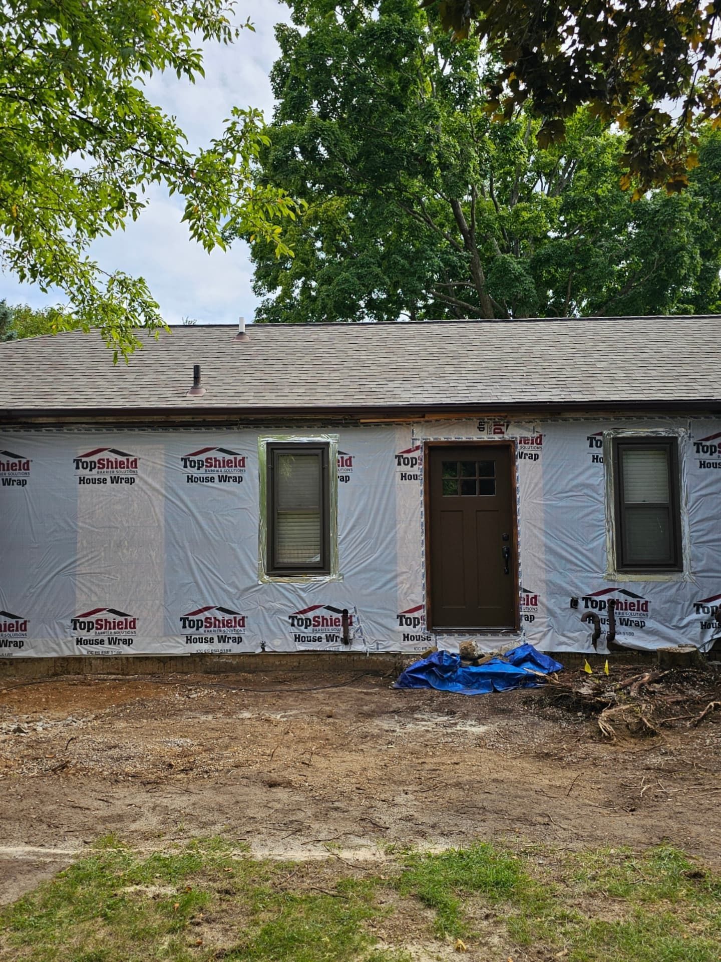 A house exterior under construction with gray house wrap, two windows, and a dark brown door, seen from a dirt yard.
