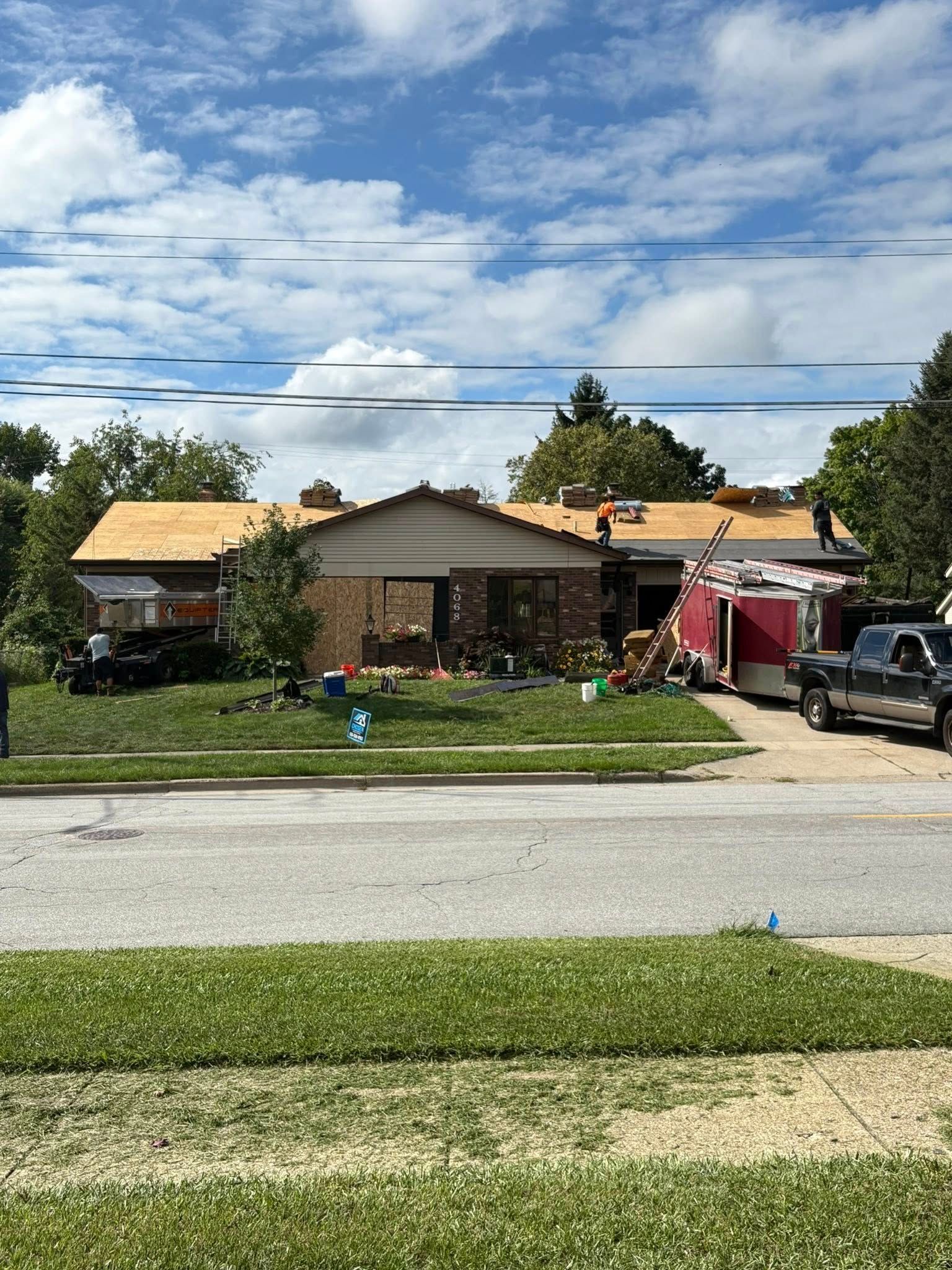 A residential house undergoing a roof replacement with workers visible on the roof and a trailer parked in the driveway.