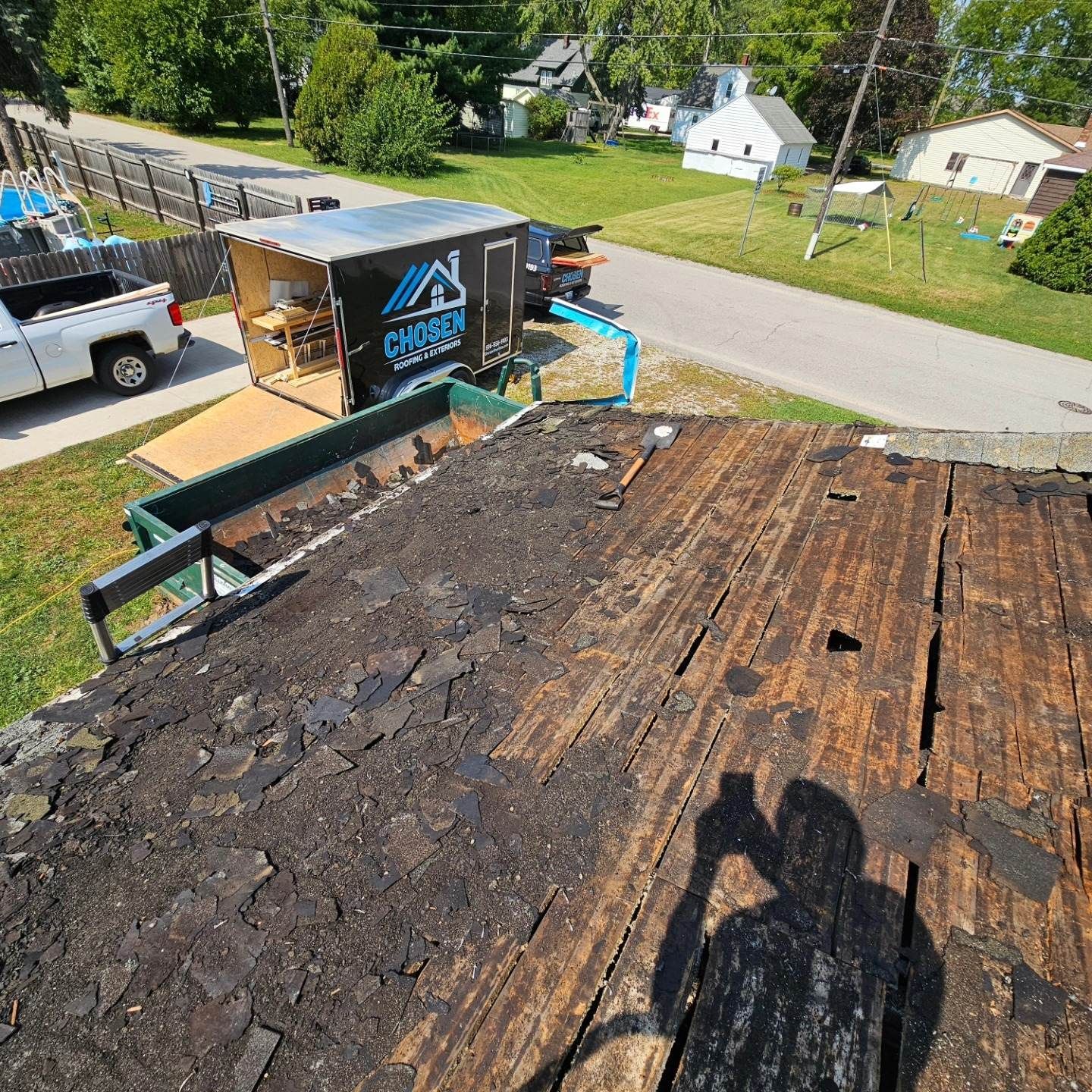 A view from a roof showing bare plywood decking under removal, a dumpster, and a work trailer on a sunny residential lot.