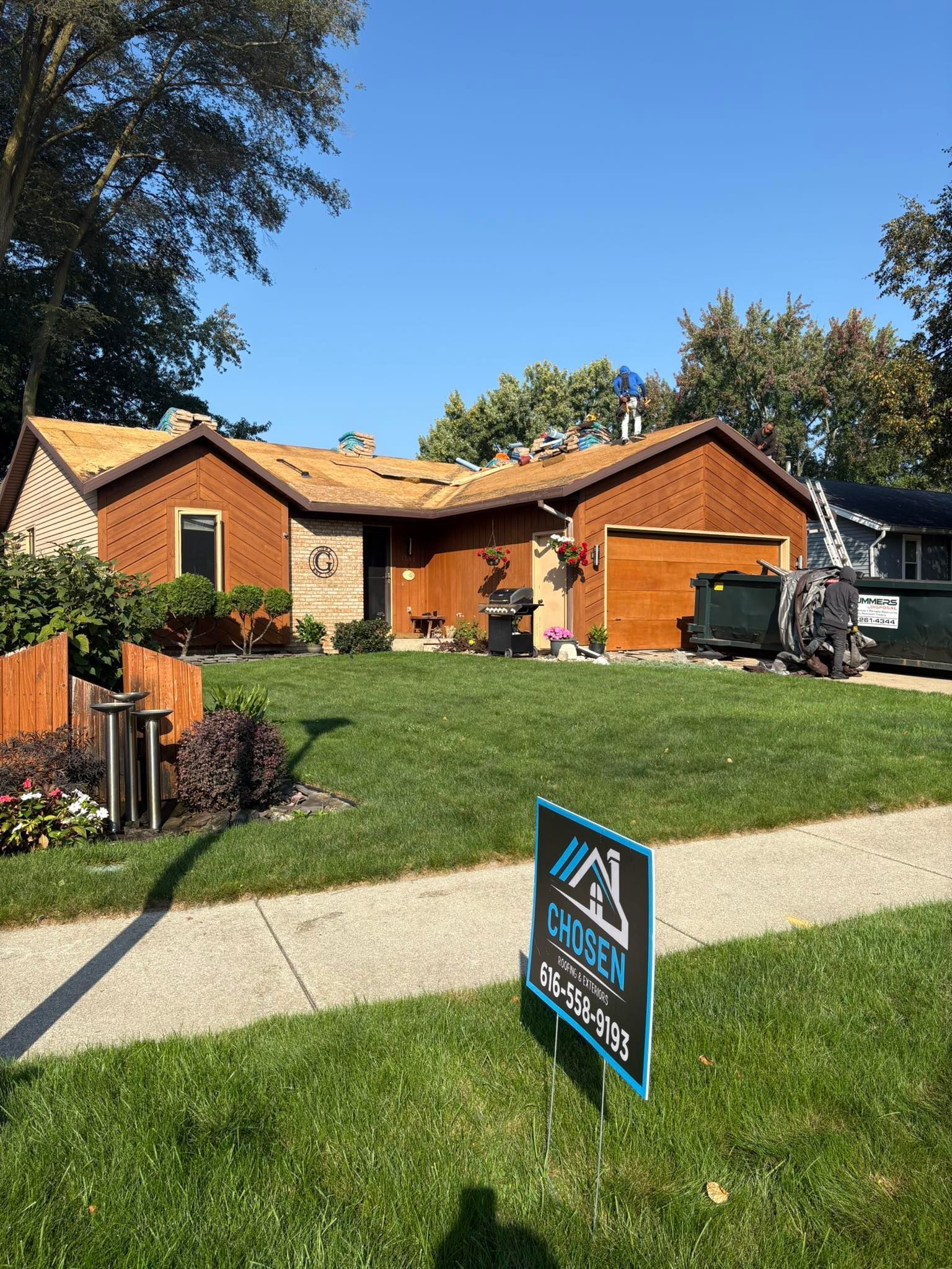 Workers replace the shingles on the roof of a brown single-story house with a lawn and a construction sign in the front.