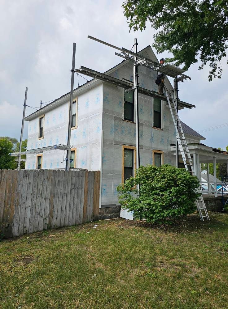 A person uses a ladder to stand on scaffolding while working on the exterior of a two-story home covered in gray wrap.