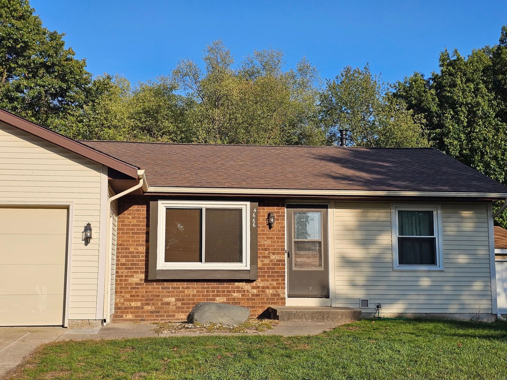 A single-story house with tan siding, a brown brick section, a white garage door, and a brown shingled roof under blue sky.