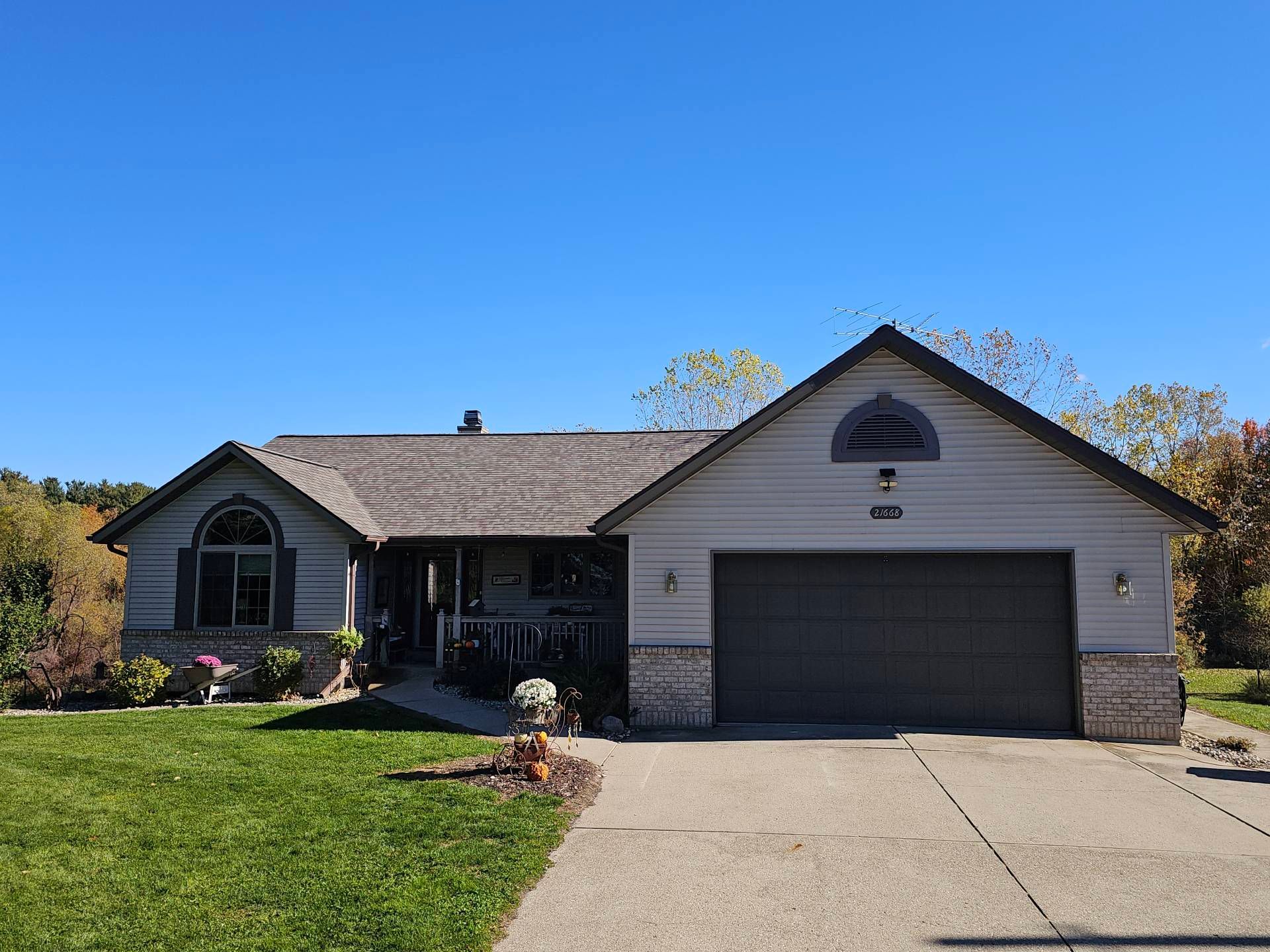 A single-story, light-colored suburban house with a two-car garage, gray roof, and a grassy front lawn under a blue sky.