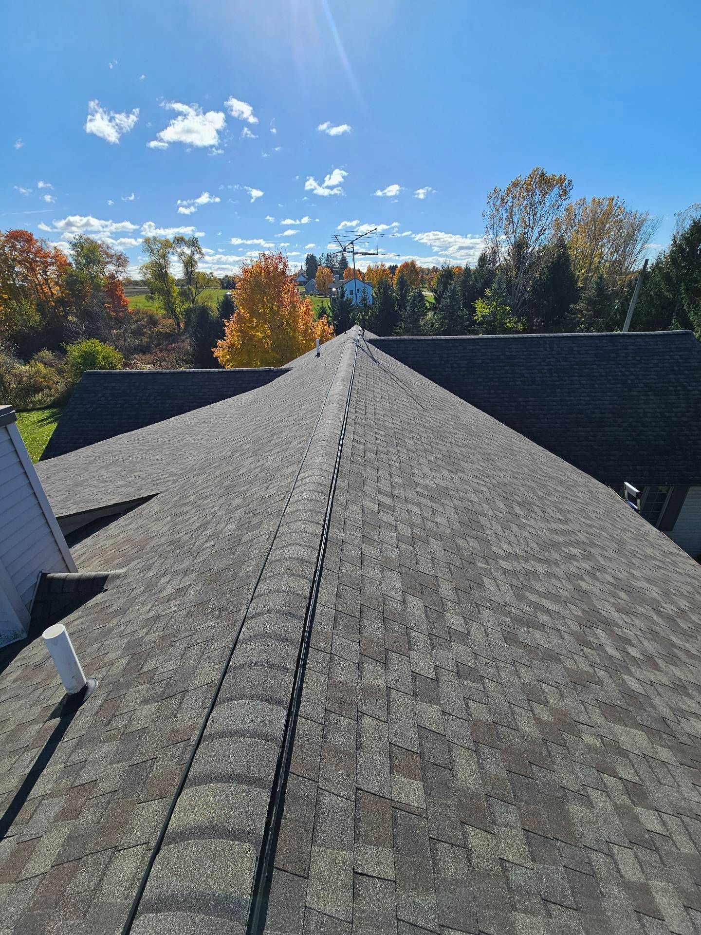 An overhead view of a dark grey shingled roof ridge extending toward a vibrant autumn forest under a clear blue sky.