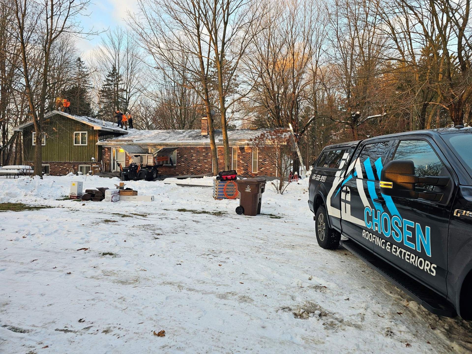 A truck from Chosen Roofing & Exteriors is parked in a snowy yard while workers replace shingles on a house roof.
