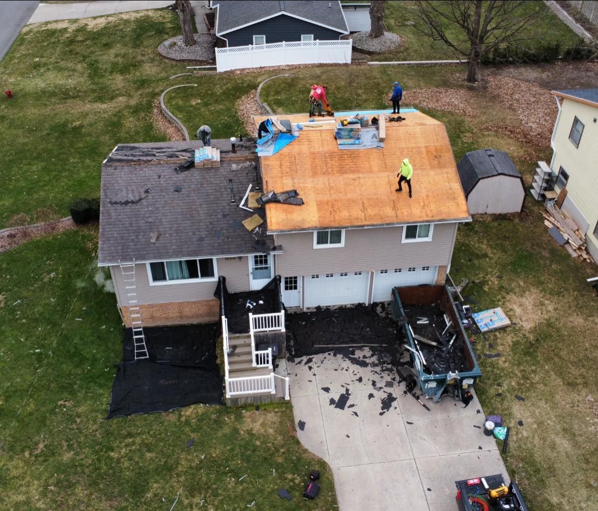 Aerial view of roofers replacing shingles on a house, with plywood exposed on one side and a dumpster in the driveway.