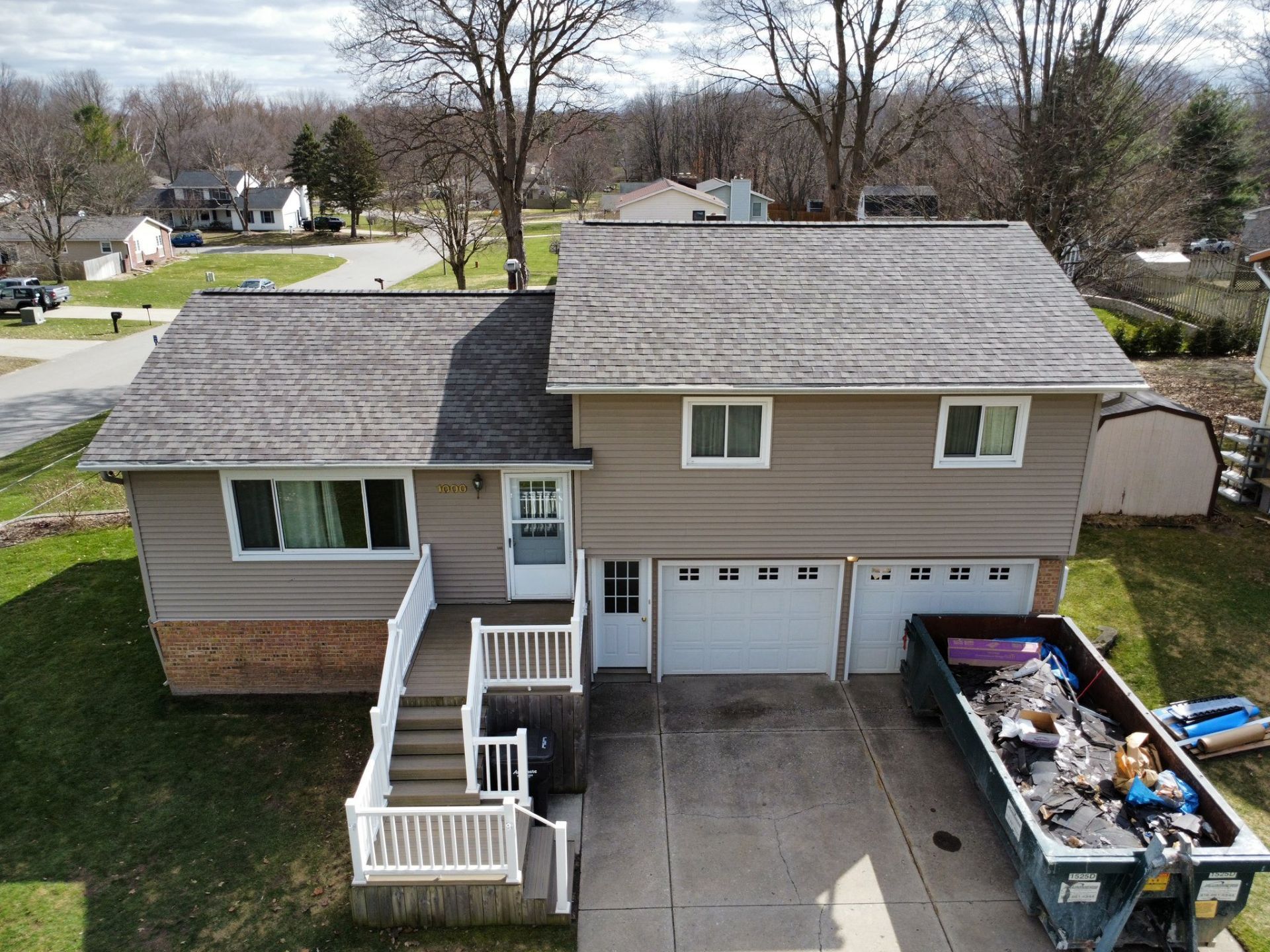 An elevated view of a beige two-story house with a grey roof, front steps, a two-car garage, and a dumpster in the driveway.