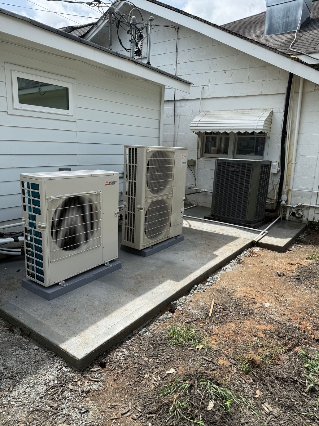 Outdoor air conditioner unit beside a brick house, with a blue hose on a concrete pad.