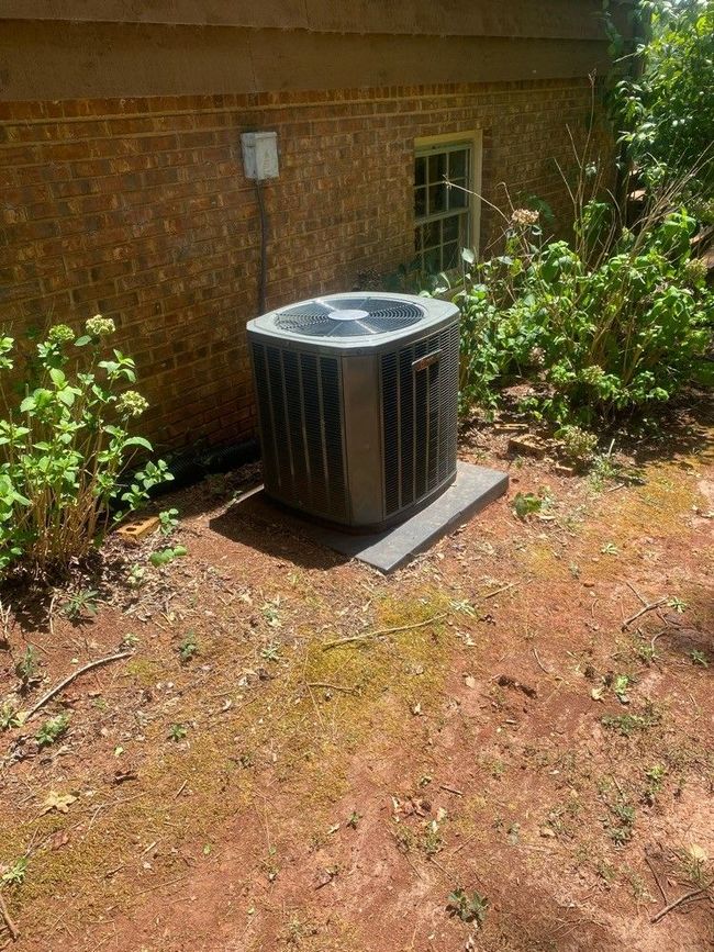 Outdoor air conditioning unit beside a brick wall, next to green bushes and a concrete pad