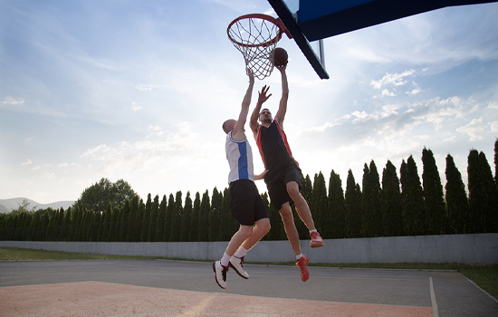 Two basketball players jump for a shot at an outdoor court at sunset.