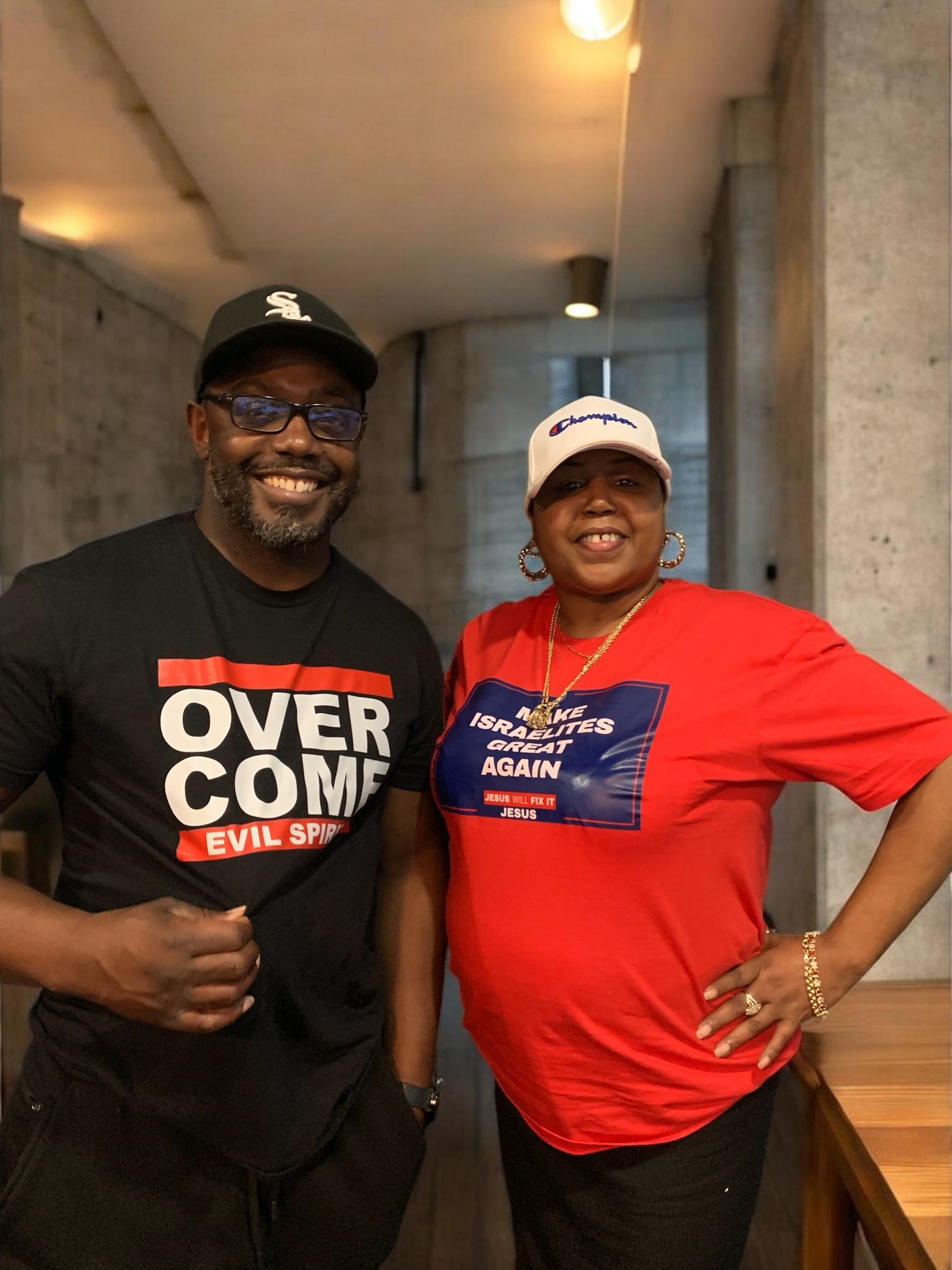 Two smiling people stand together in a hallway, wearing black and red T-shirts and baseball caps.