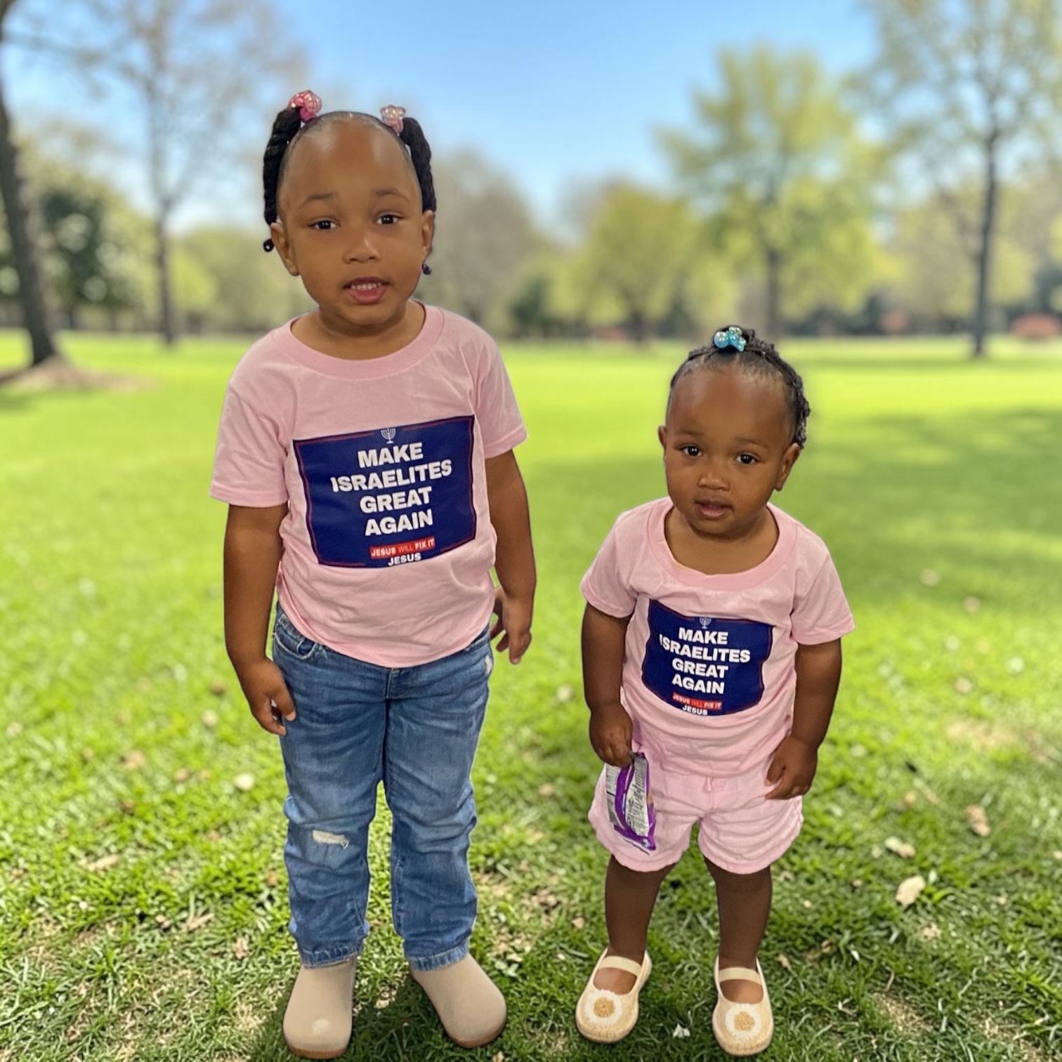 Two children in matching pink shirts standing on grass in a sunny park