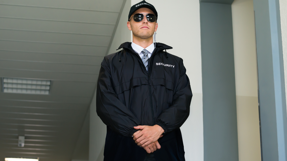 Security guard in black uniform, sunglasses, and cap, standing with arms crossed.