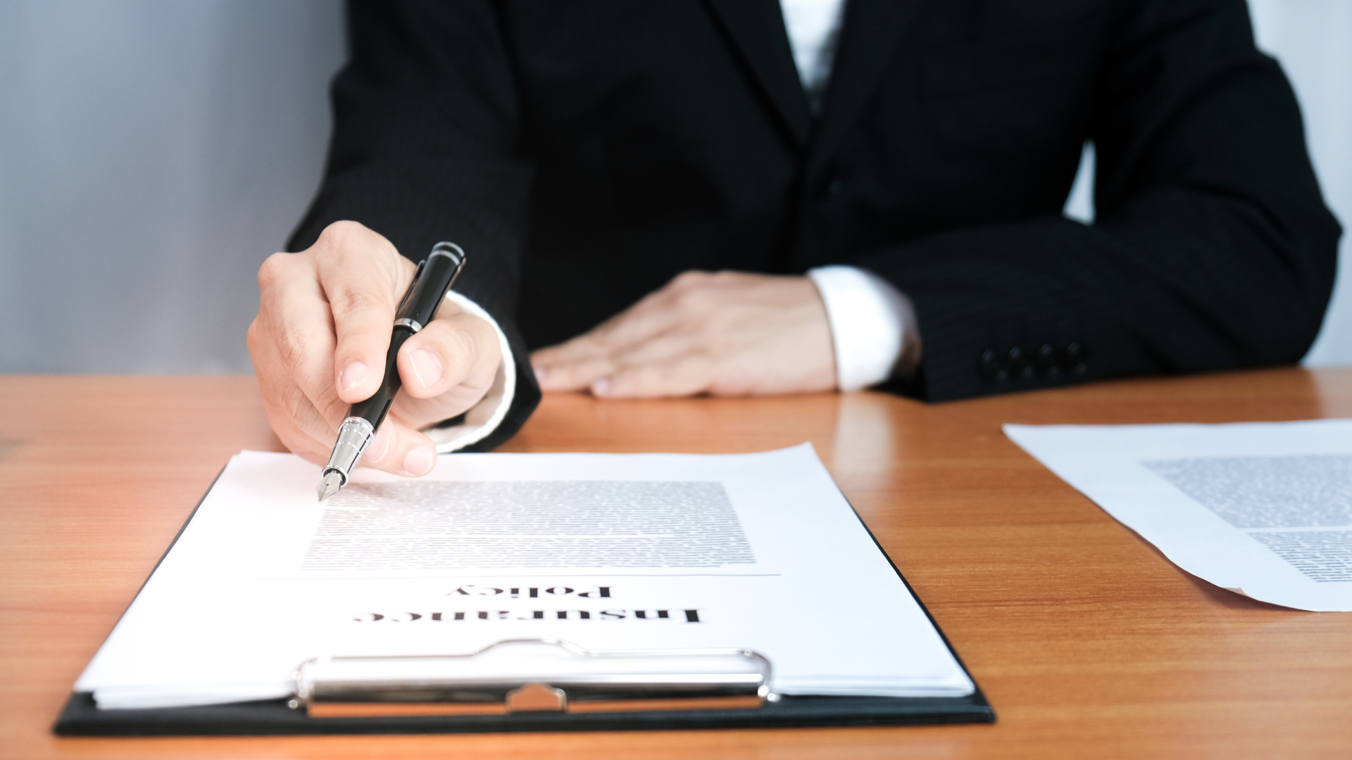 Person in suit signing a document on a clipboard at a wooden desk.