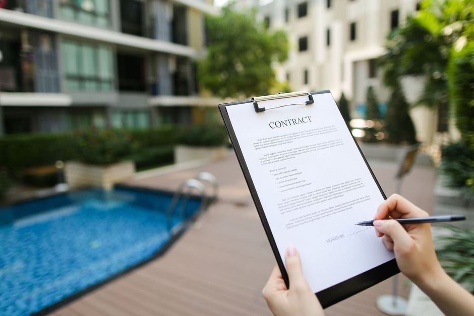 Person holding contract in front of a pool and condos, signing with pen.