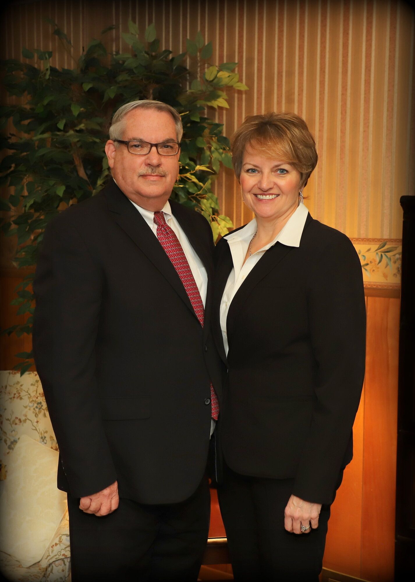 Two people in black formalwear standing indoors, smiling in front of striped curtains and a leafy plant.