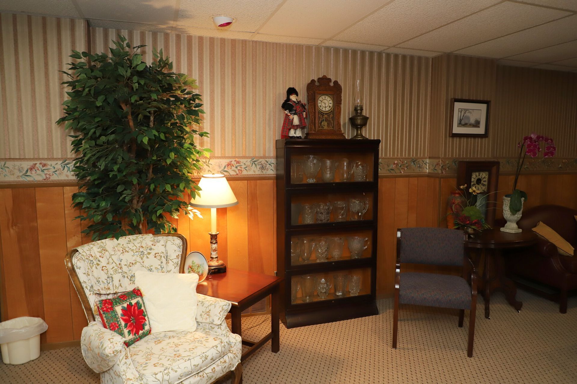 Cozy waiting room with armchairs, lamp, bookshelf, and potted plant against wood-paneled walls.