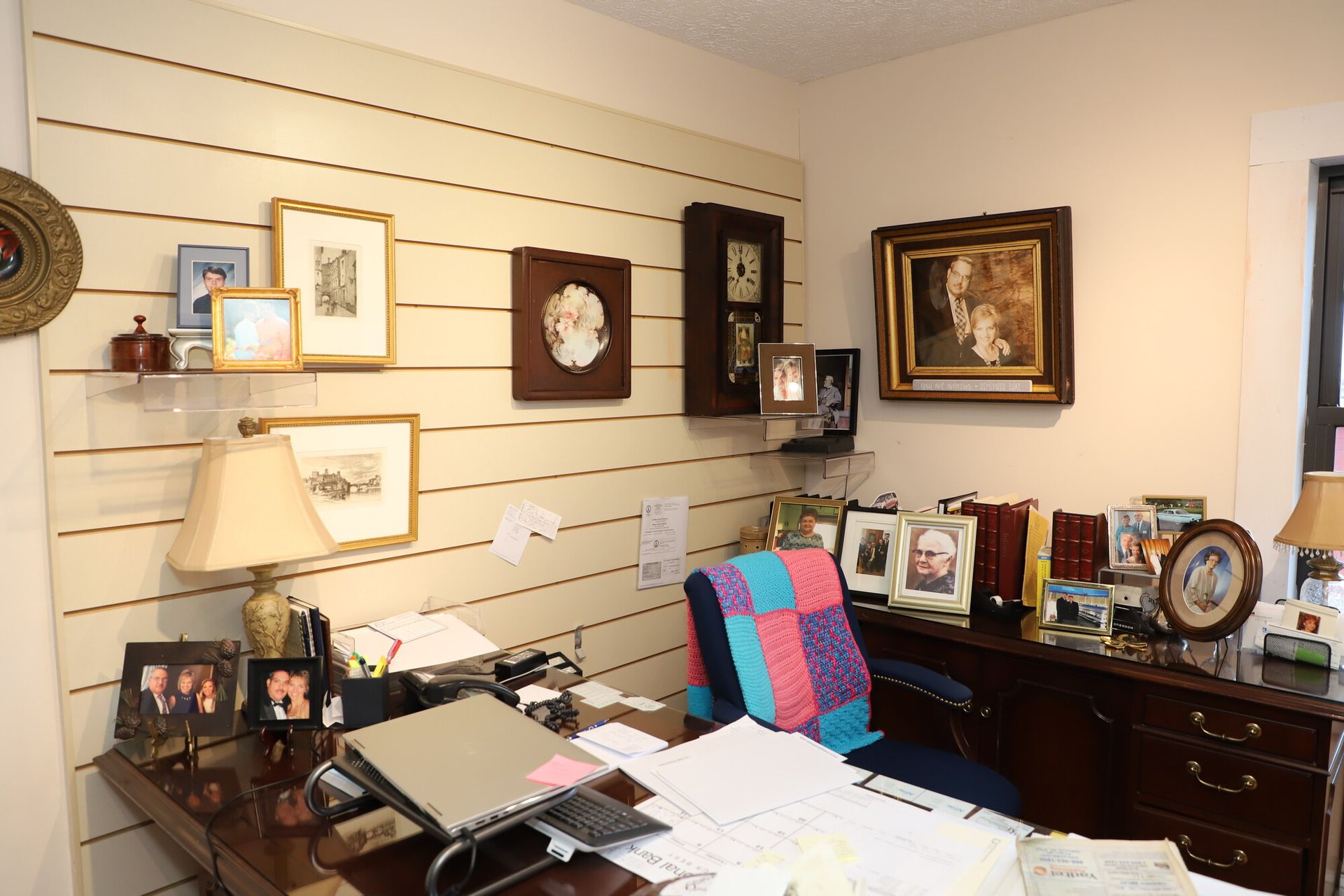 Home office with desk, framed photos, wall art, and a colorful quilt draped over a chair.
