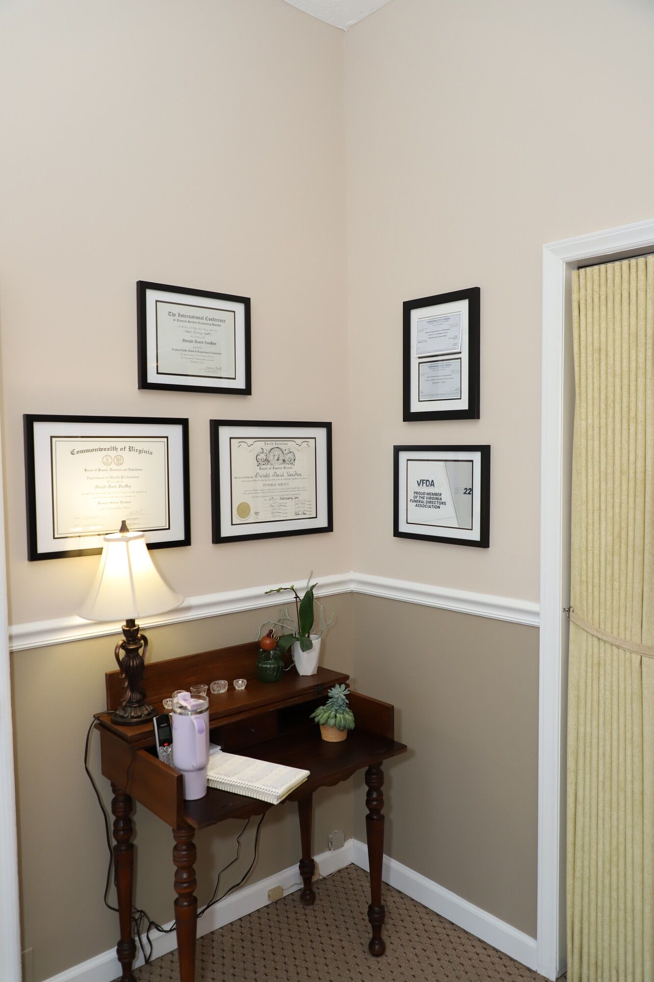 Corner office nook with framed certificates, a small wooden desk, and a lamp against beige walls.