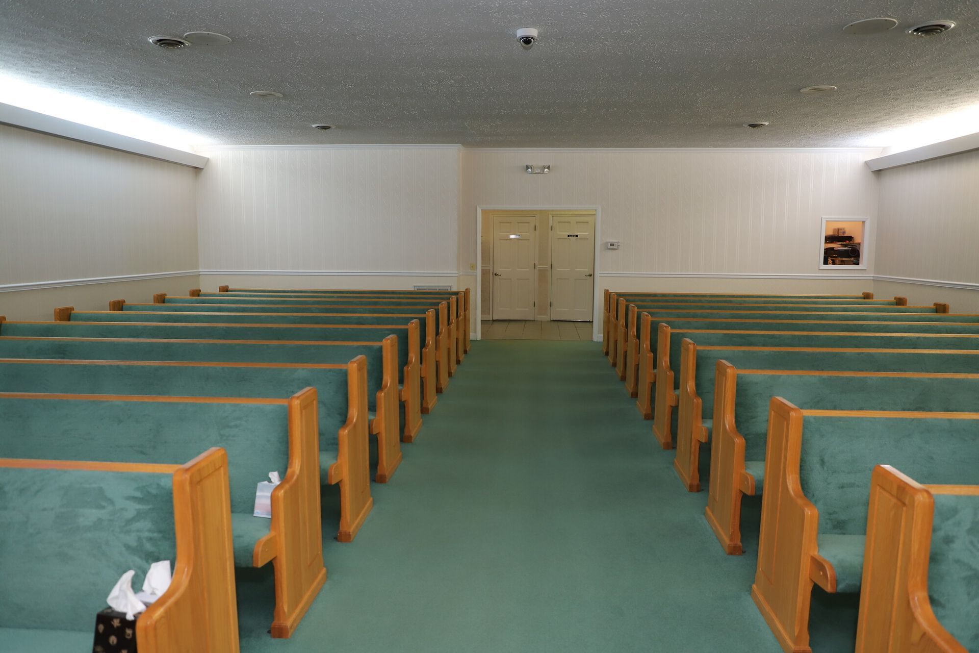 Empty church sanctuary with green carpeted pews facing double doors at the front.