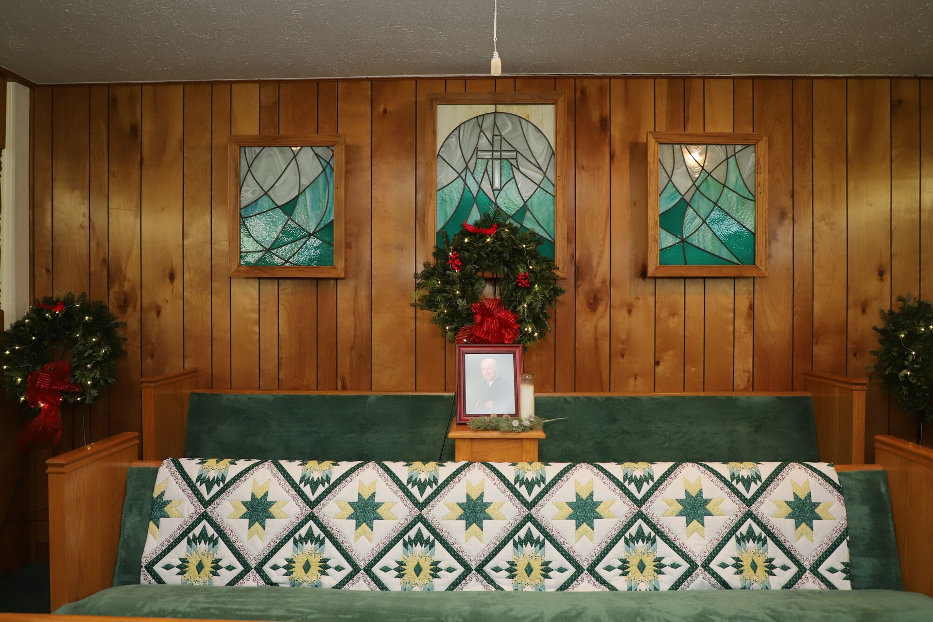 Church altar with green cloth, stained-glass windows, and Christmas wreaths on wood-paneled wall