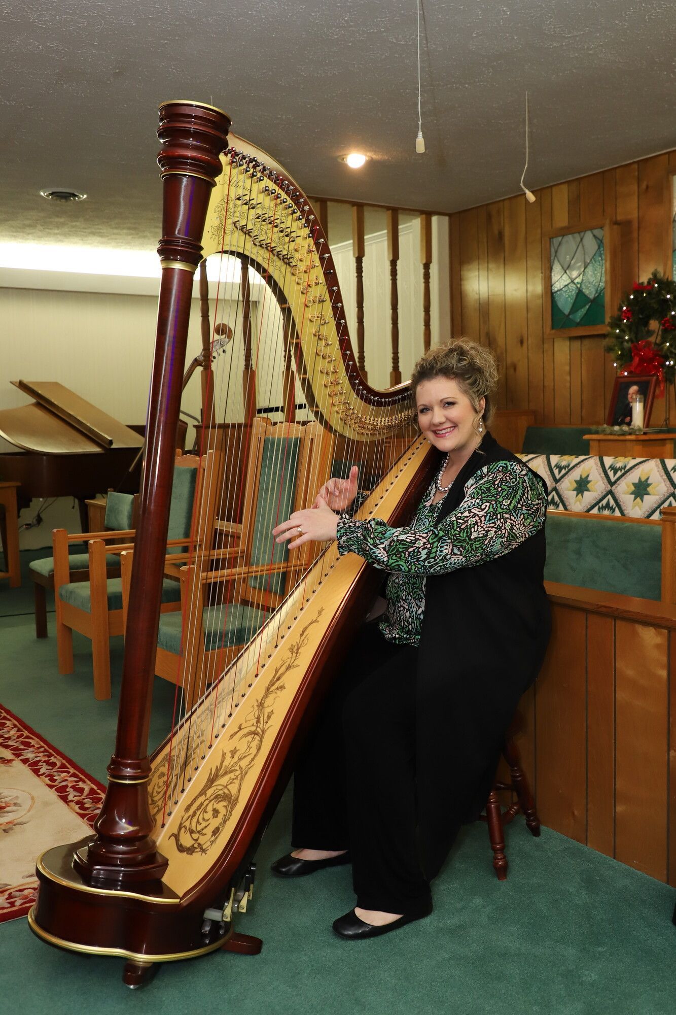 Woman seated playing a gold harp in a church-like interior with wooden chairs and green carpet