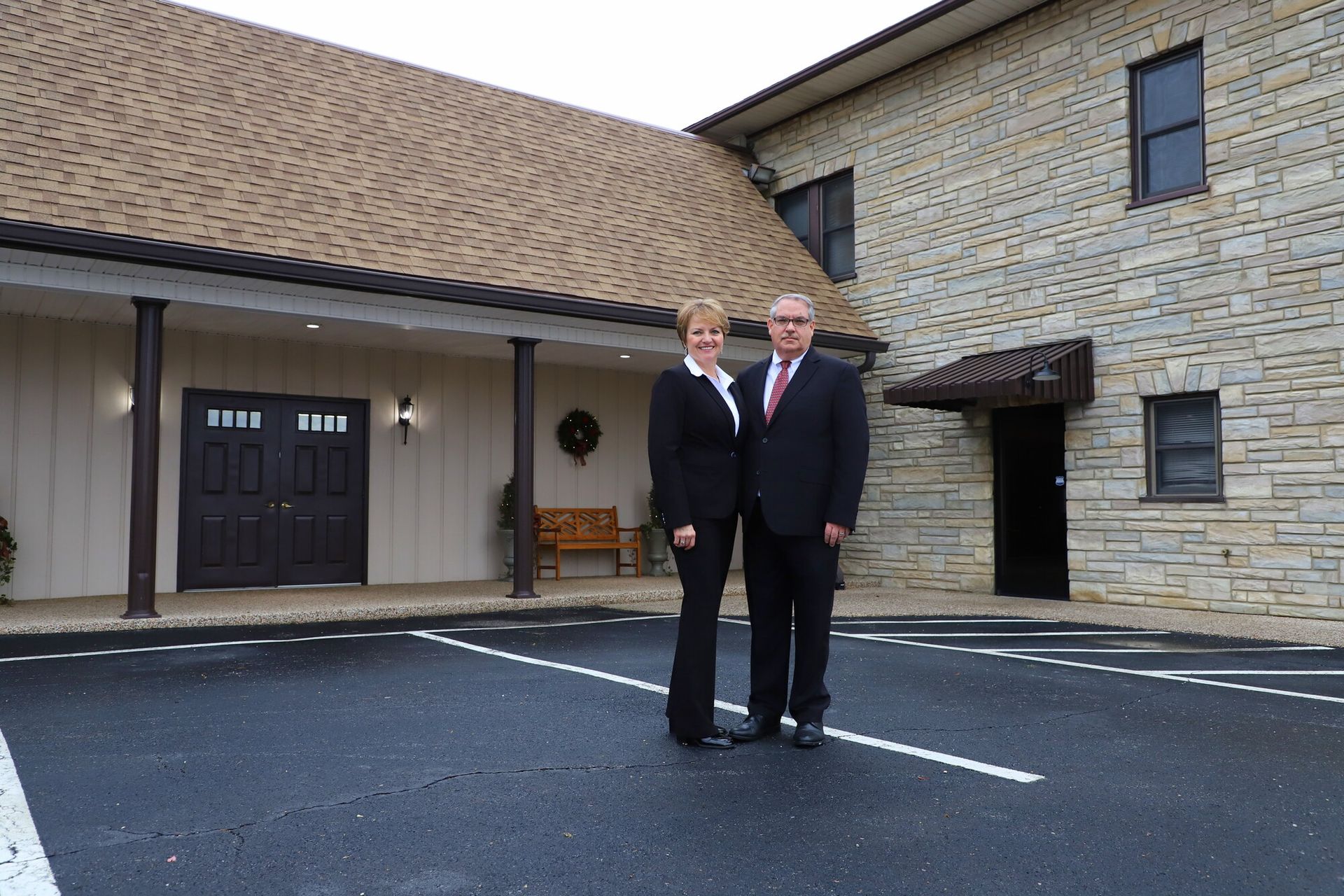 Two people in suits standing outside a stone building with a dark doorway and parking lot.