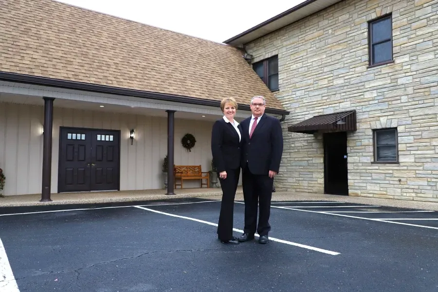 Two people standing in a parking lot outside a stone building with a covered entrance